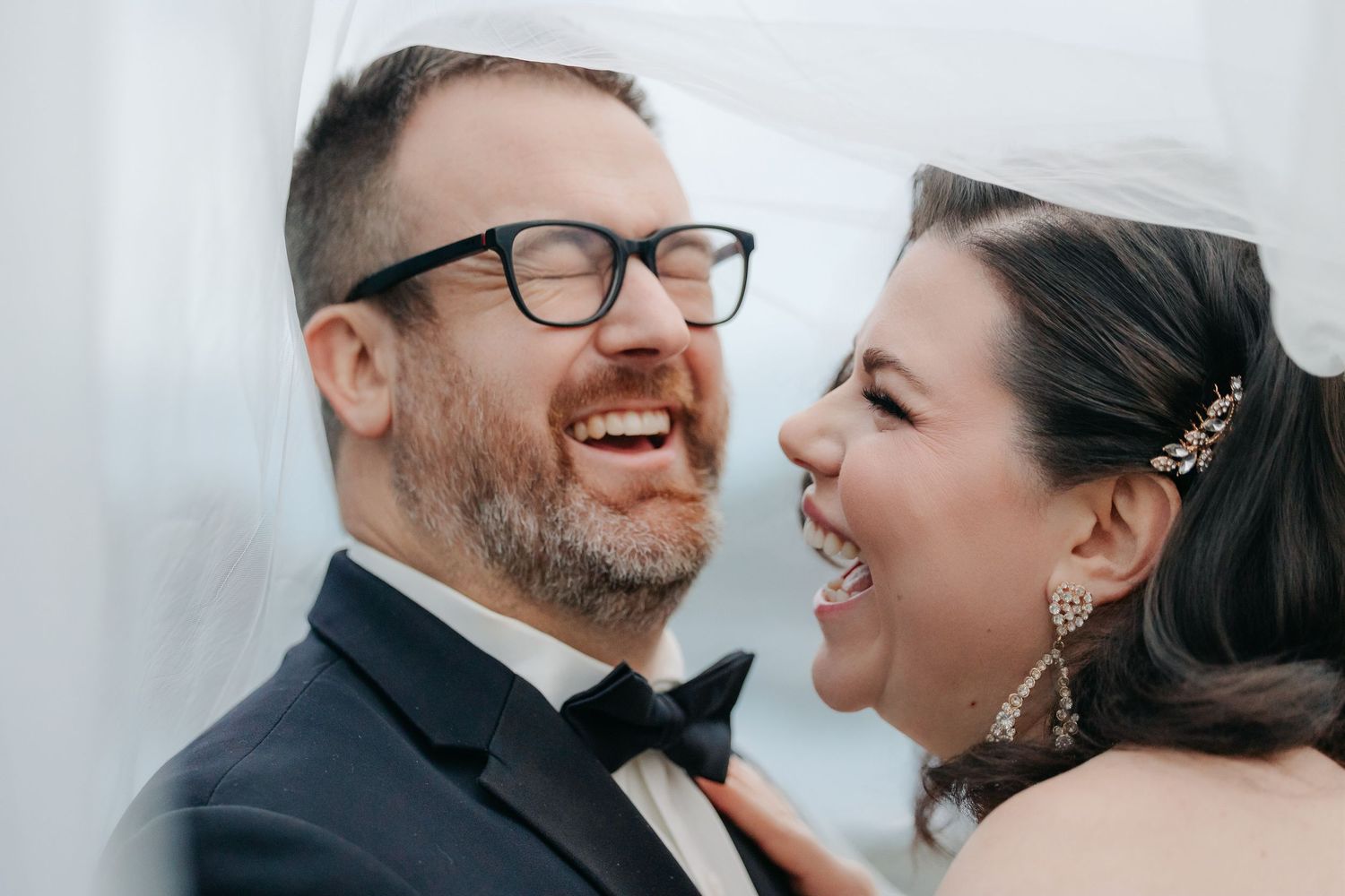 Laughing bride and groom sharing a joyful moment, groom wearing glasses and bow tie on their wedding day.