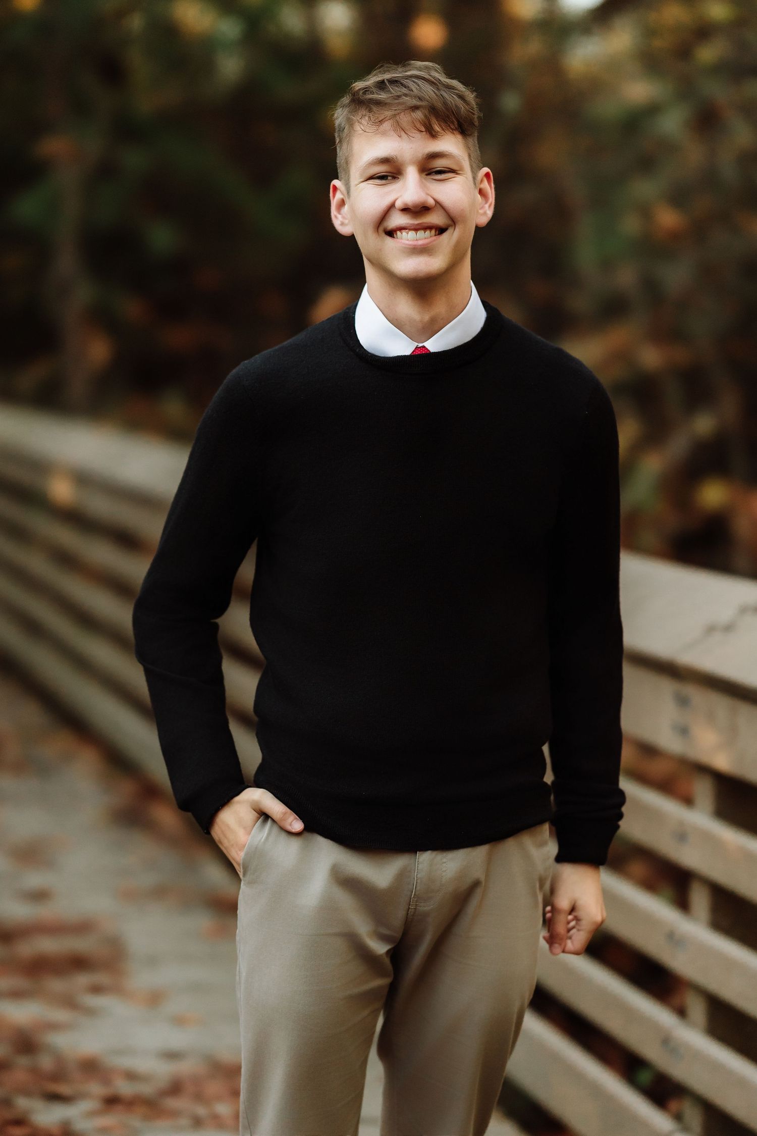 Smiling young man in black sweater and khaki pants standing on a wooden bridge surrounded by autumn foliage.
