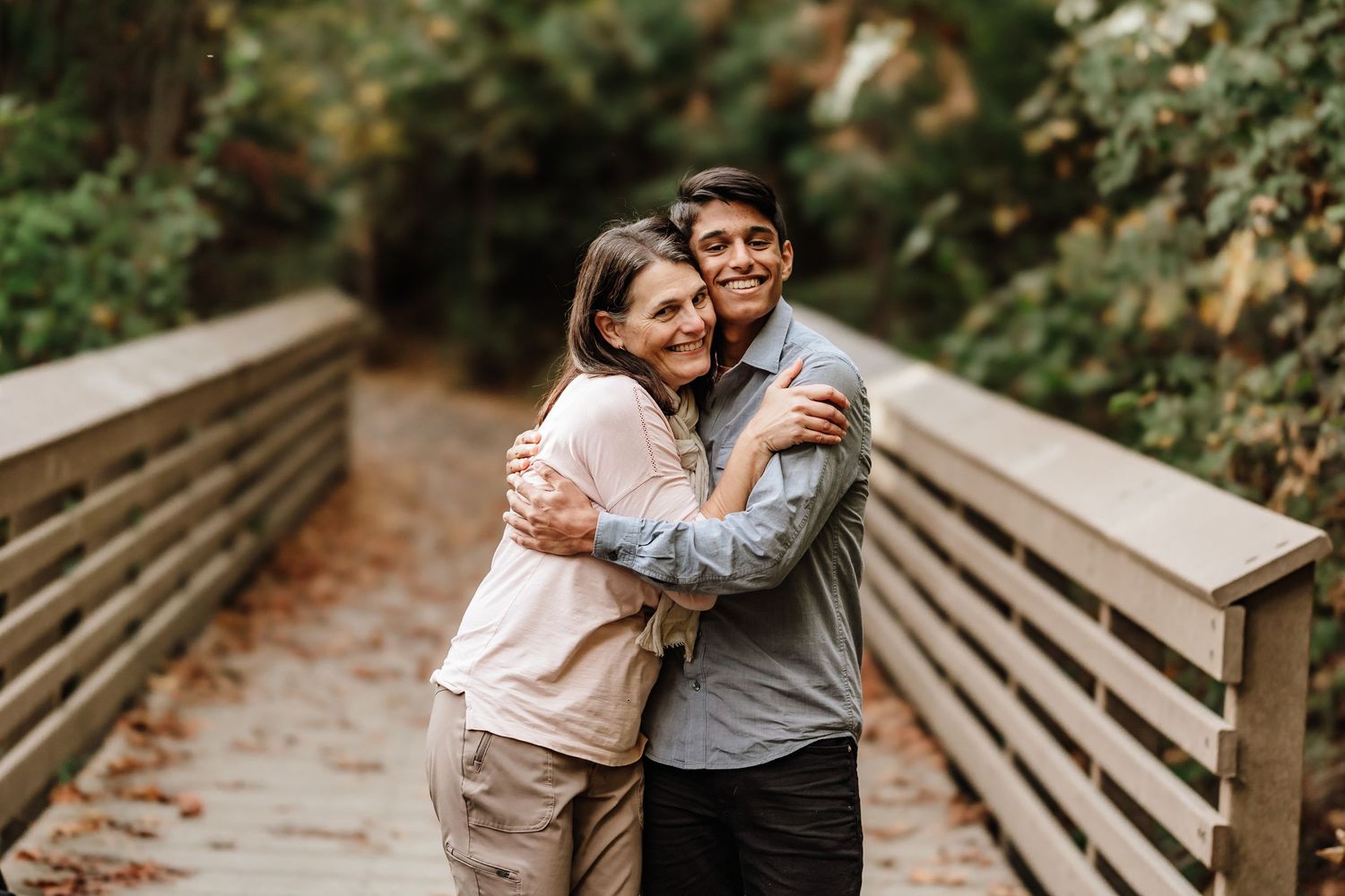 A smiling couple embraces warmly on a wooden bridge surrounded by lush green trees in a park setting.