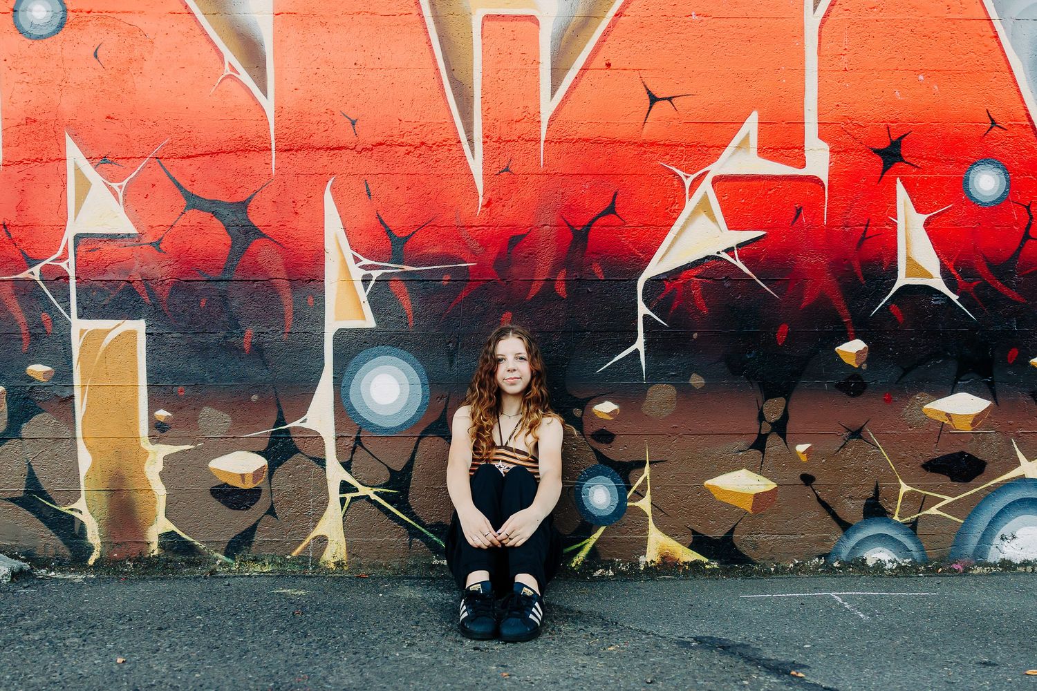 Young woman with red hair sitting against a vibrant graffiti mural wall in red and gold tones.