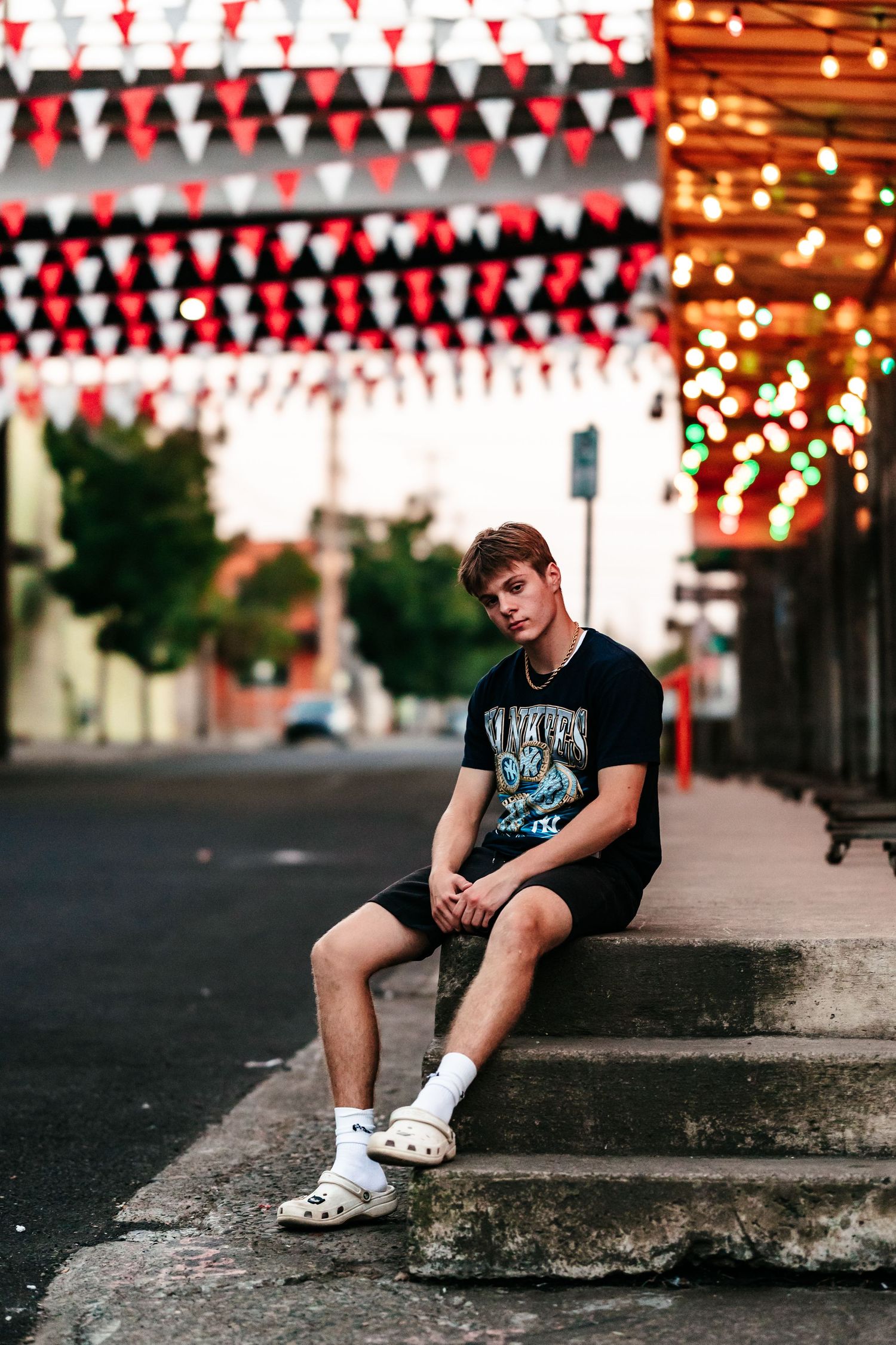 Young man in black graphic tee and Crocs sits on steps under colorful string lights and triangle bunting flags.