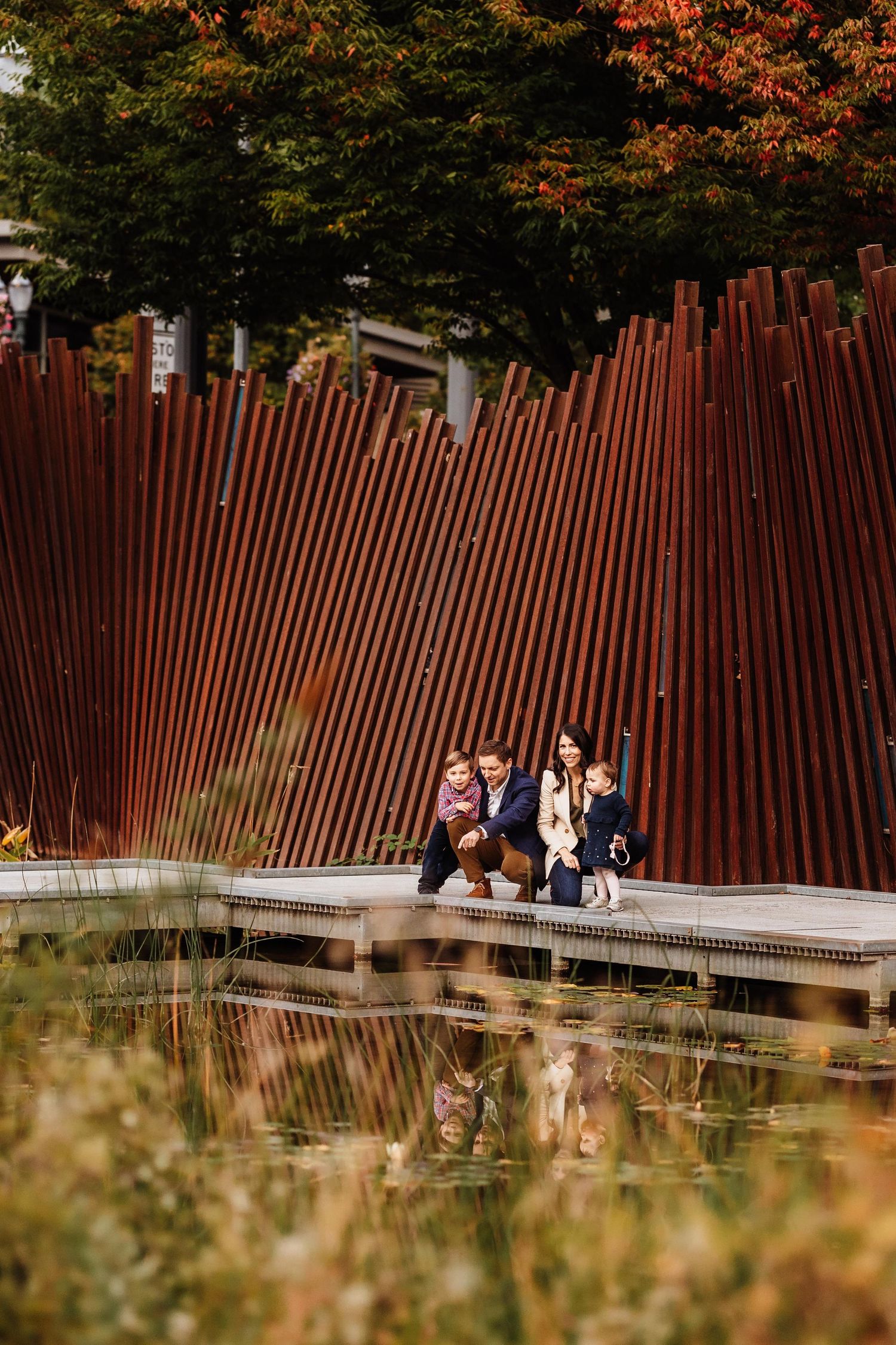 Family of four poses by a rustic metal fence near a reflective pond surrounded by autumn foliage.