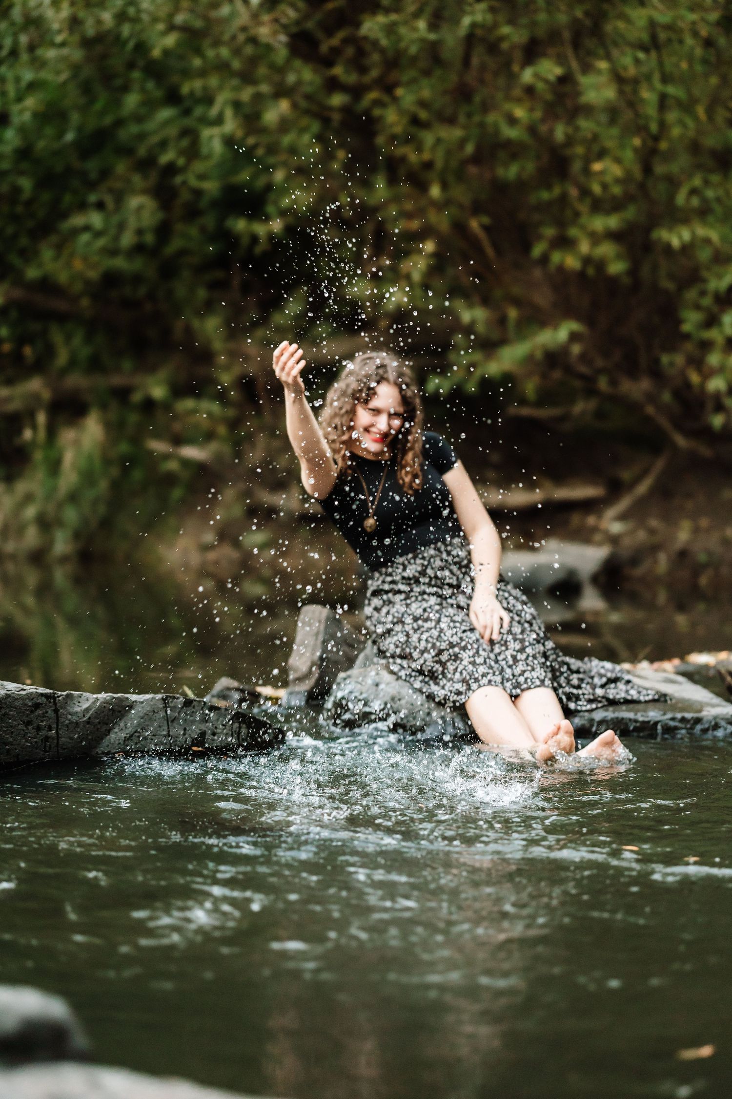 Happy woman in floral dress splashing water with her feet while sitting on rocks in a forest creek.