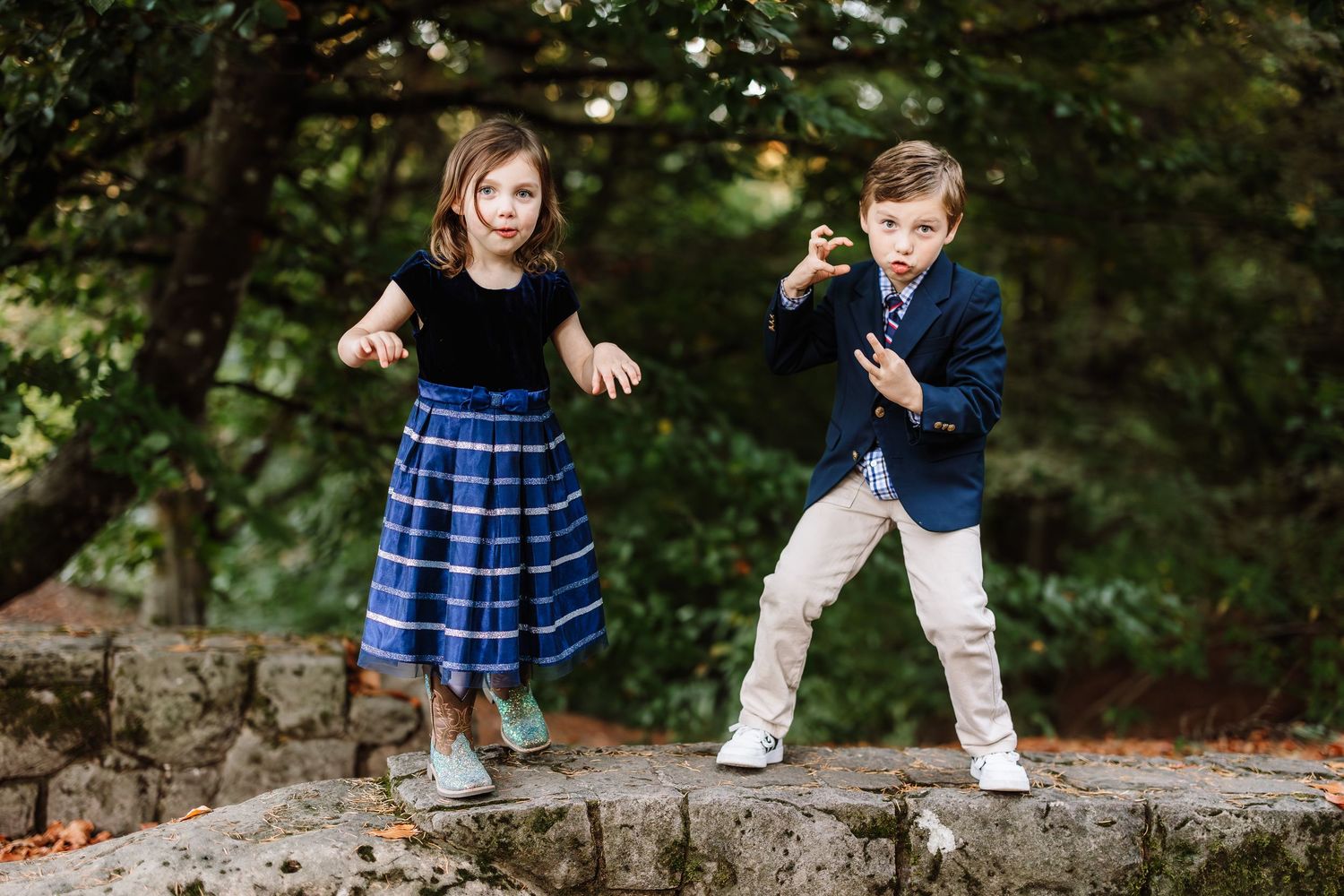 Two children posing outdoors on stone wall, girl in blue dress and boy in navy blazer, surrounded by lush green trees.