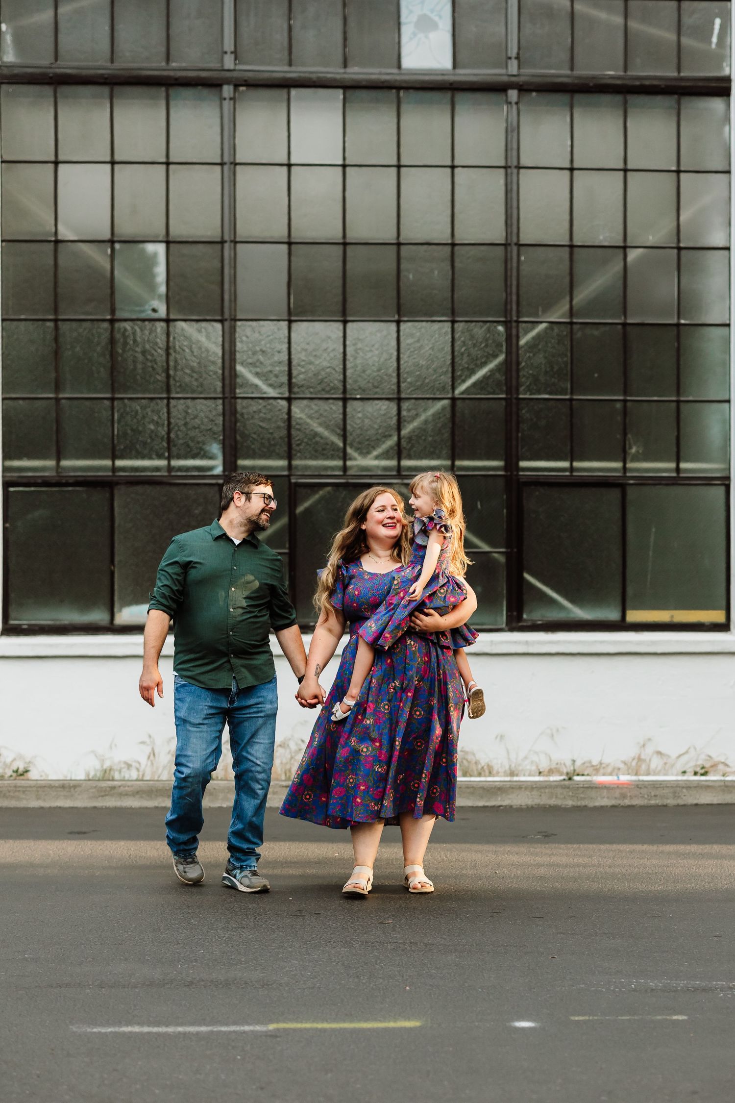 A happy family of three walks outdoors, mom in purple floral dress holds child, dad in green shirt, near industrial windows.