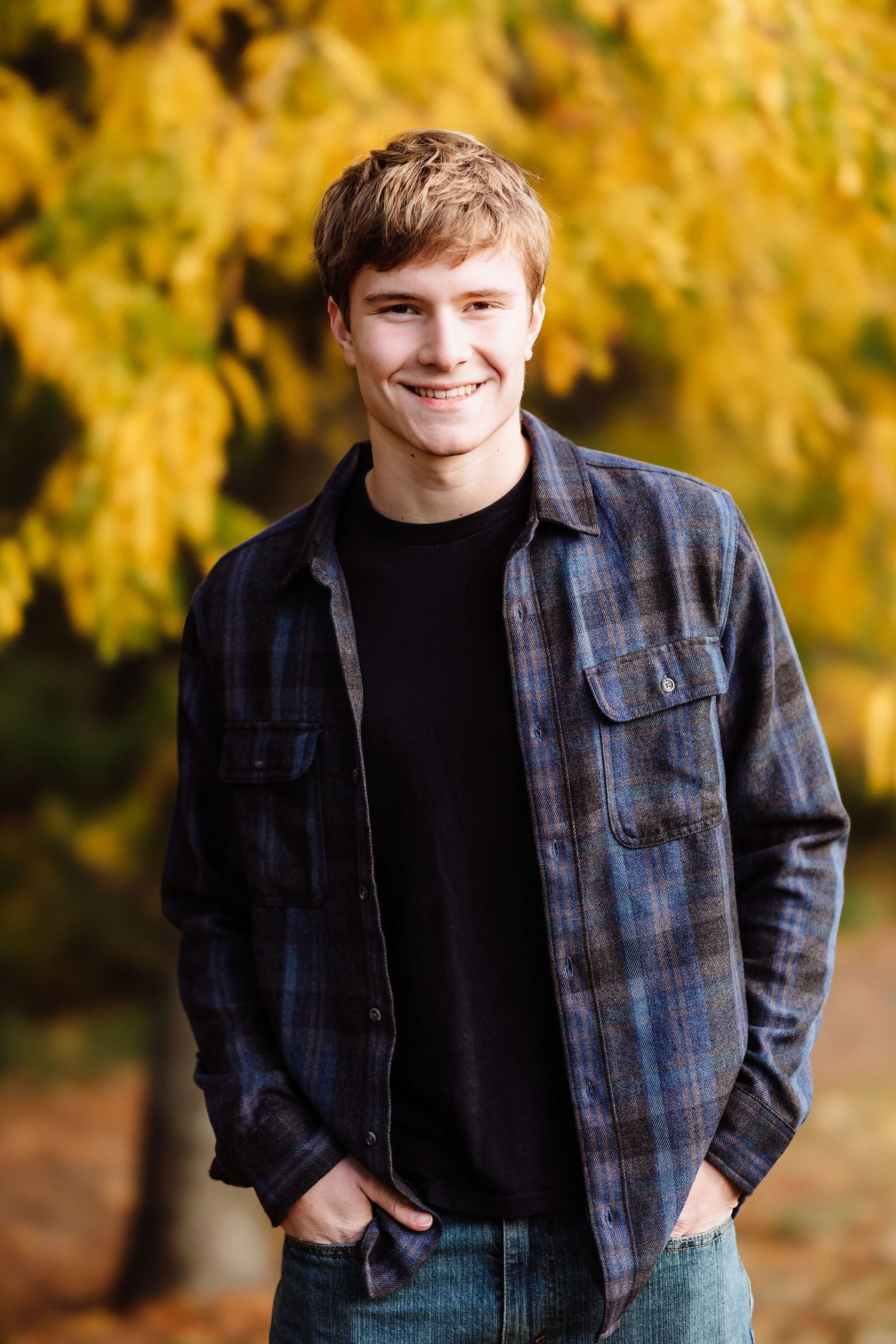 Smiling young man in plaid shirt and jeans poses outdoors with vibrant yellow autumn foliage in background.