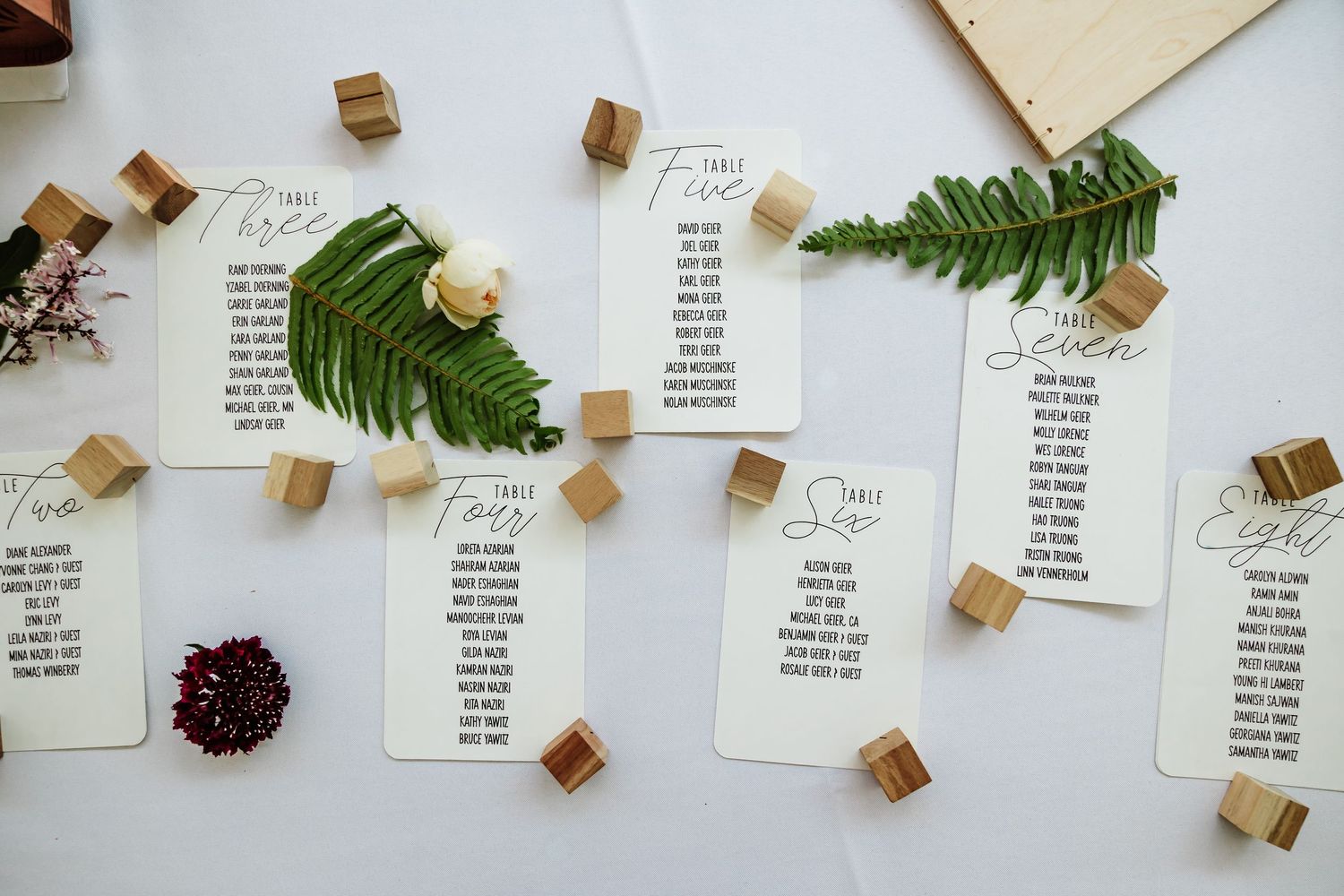 Wedding seating chart cards displayed on wooden cube holders with fern and floral decorations on white surface.
