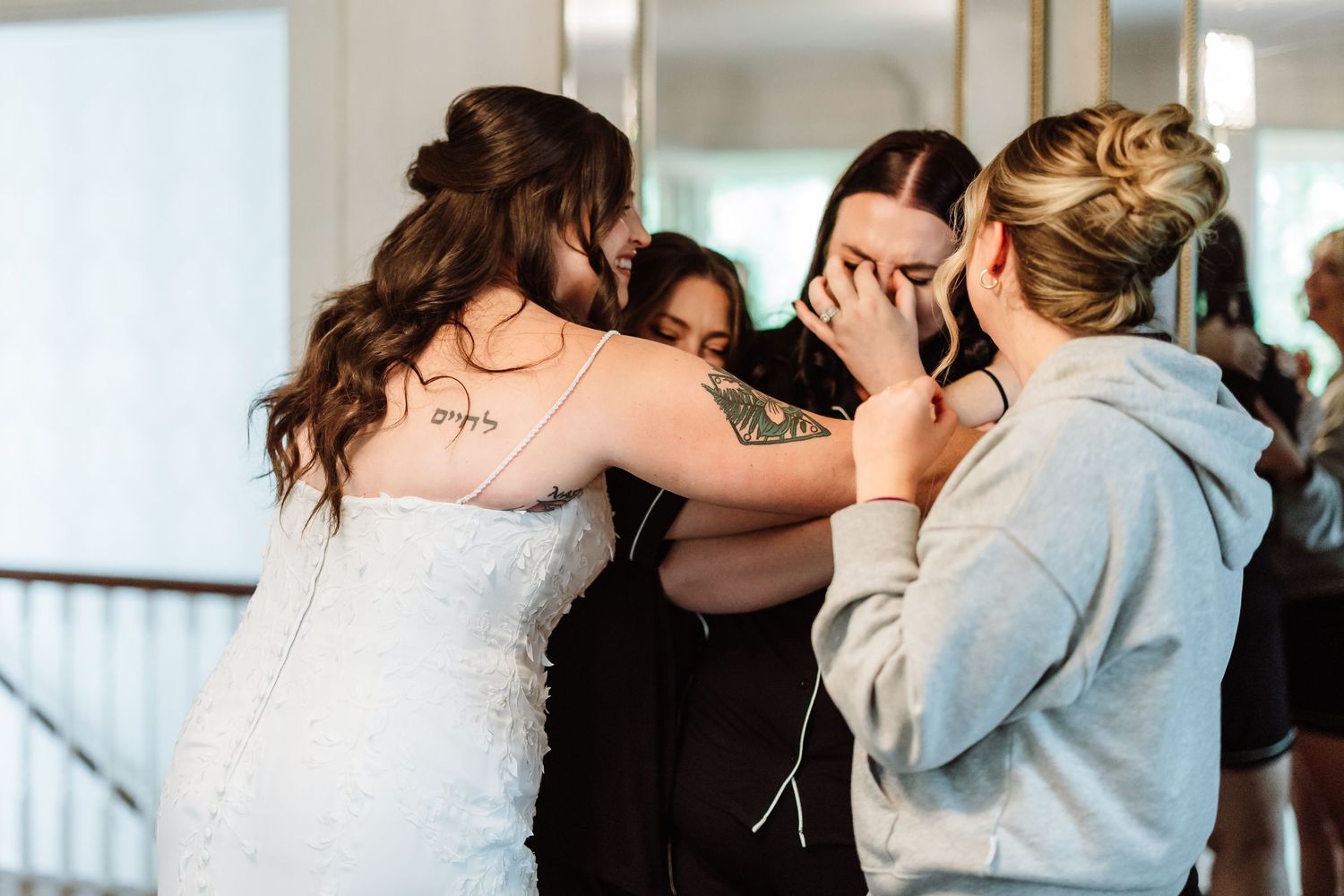 Emotional bride in lace gown sharing a group hug with tearful bridesmaids.
