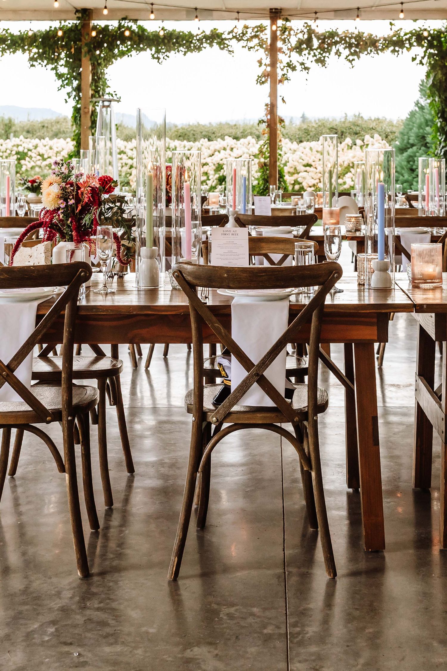 Rustic wedding reception with wooden farm tables, cross-back chairs, floral centerpieces, and tall candles under a draped tent.