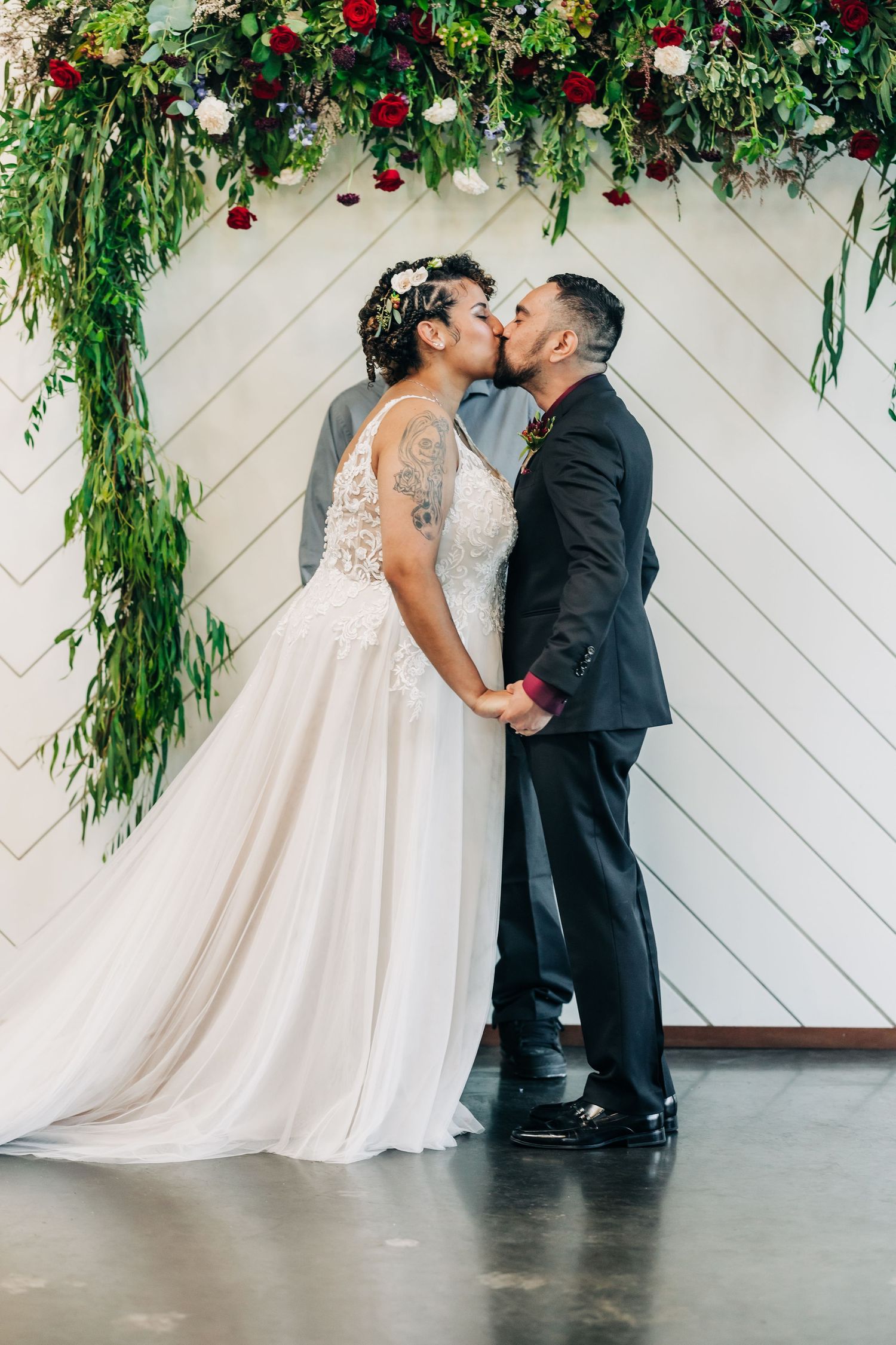 Bride and groom share first kiss under floral arch at indoor wedding ceremony.