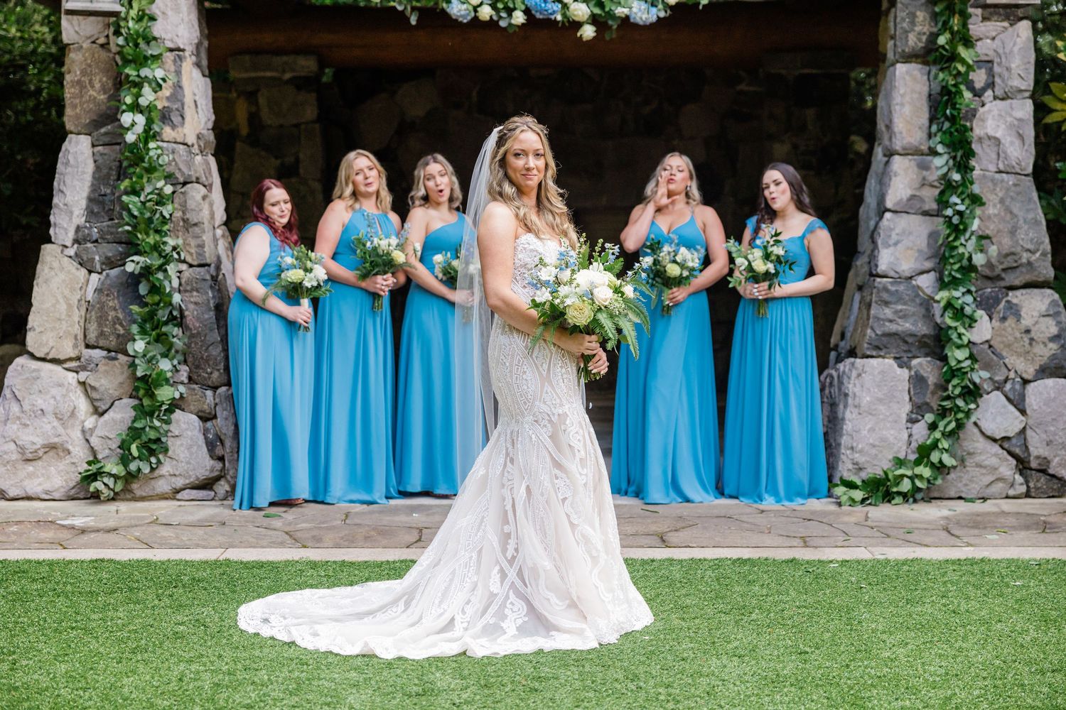 Bride in lace gown with veil stands before bridesmaids in blue dresses at stone chapel.