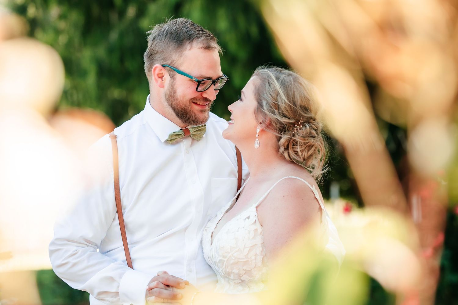 Bride and groom gazing at each other outdoors, groom wearing suspenders and a colorful bow tie in golden light.