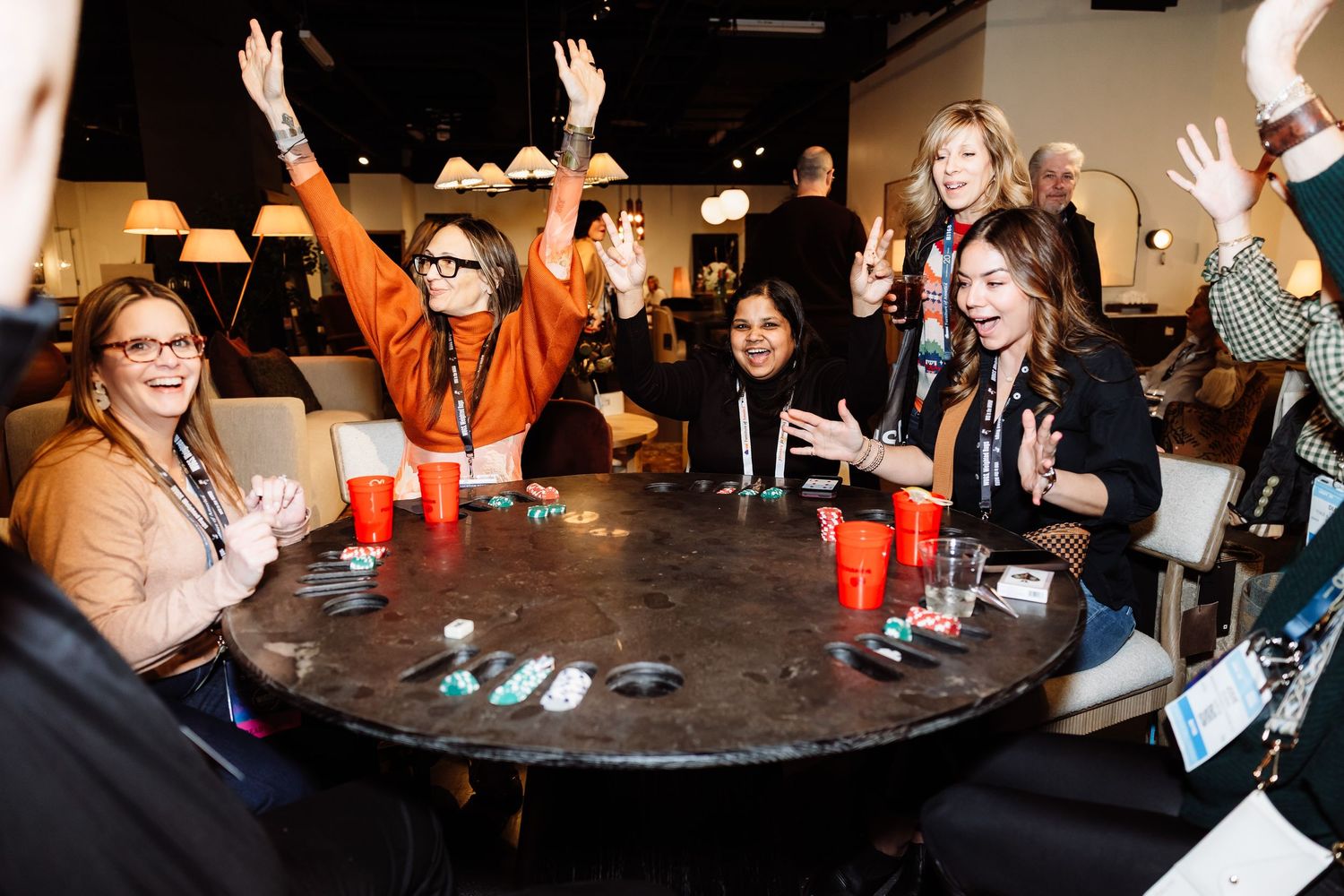 A group of smiling women celebrate with raised hands around a round table at an indoor event.