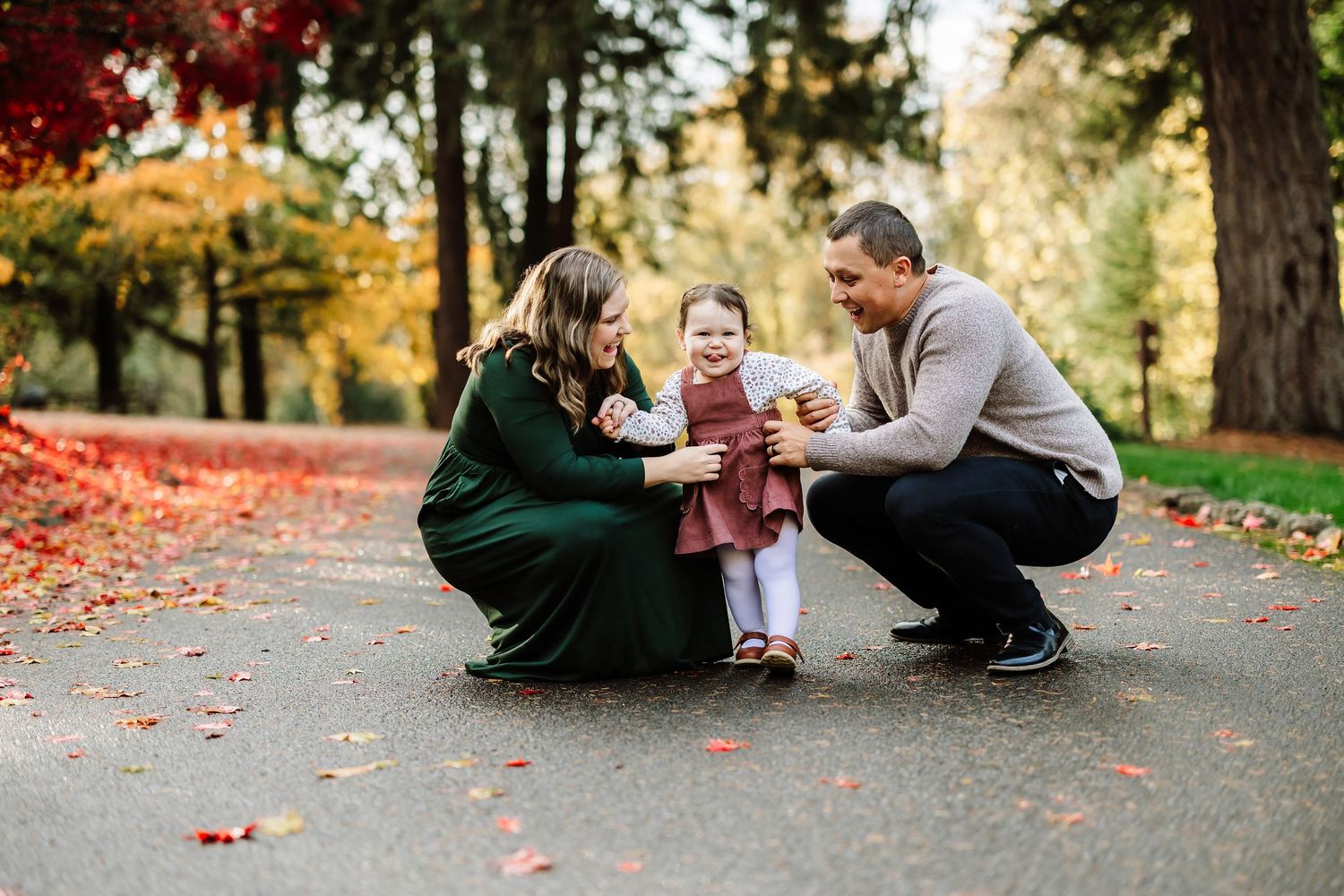 Family of three enjoying autumn park, parents crouching beside toddler girl in pink dress among colorful fallen leaves.