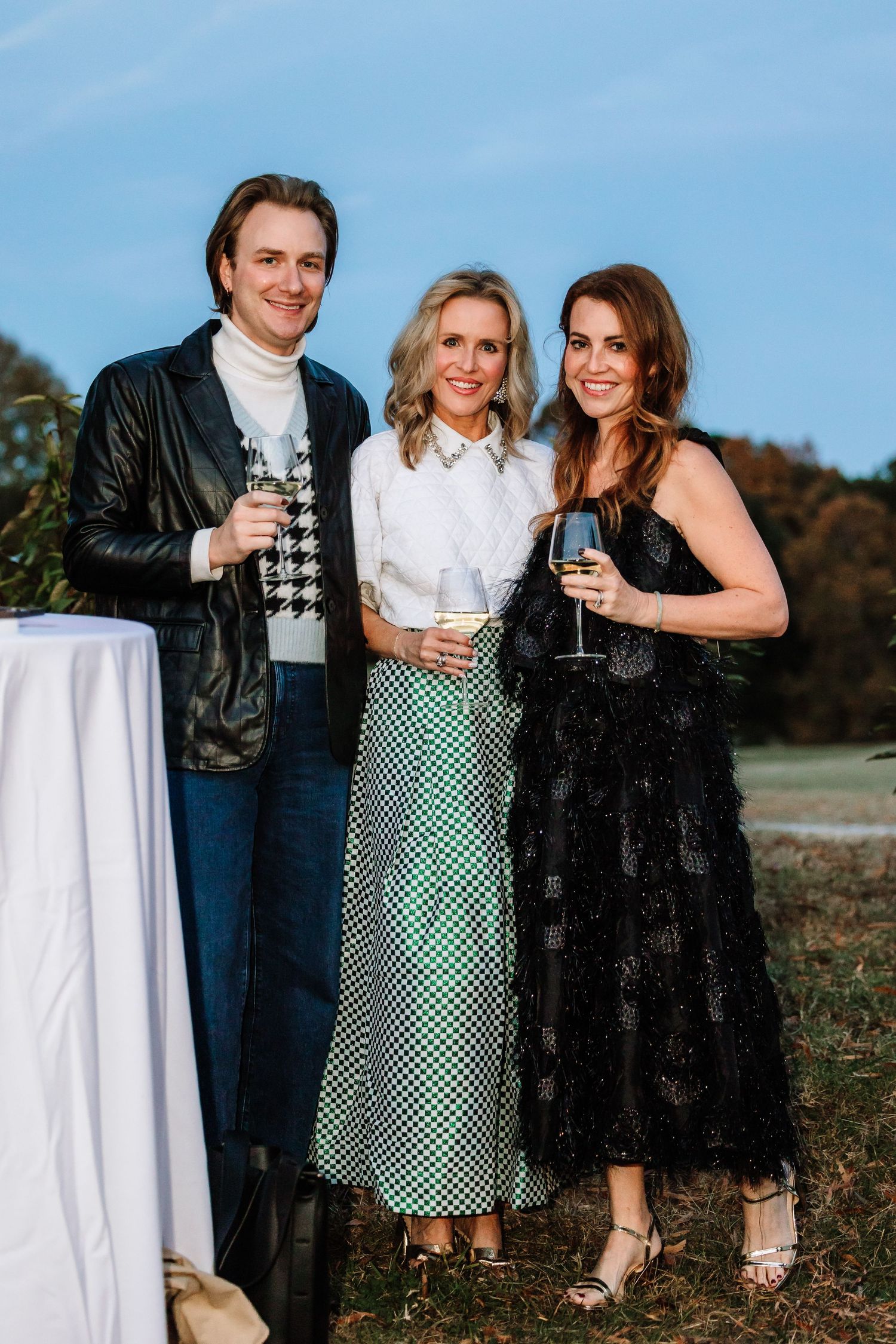 Three elegantly dressed guests holding champagne glasses at an outdoor evening social event.