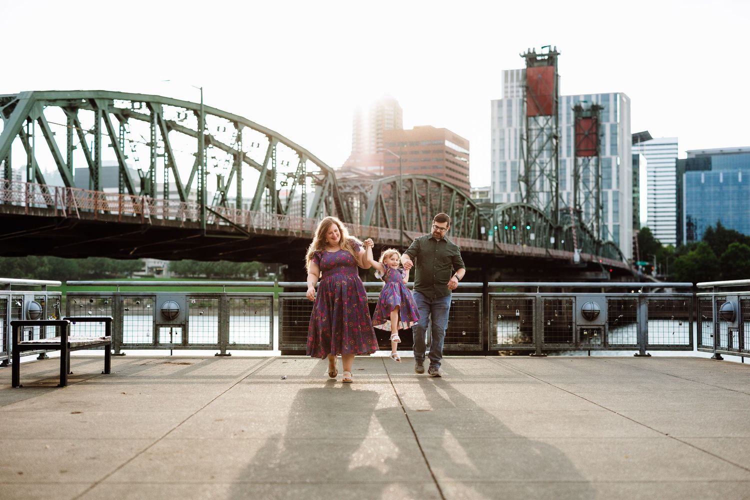 Family of three holding hands on a waterfront plaza with a green arch bridge and city skyline in Portland, Oregon.