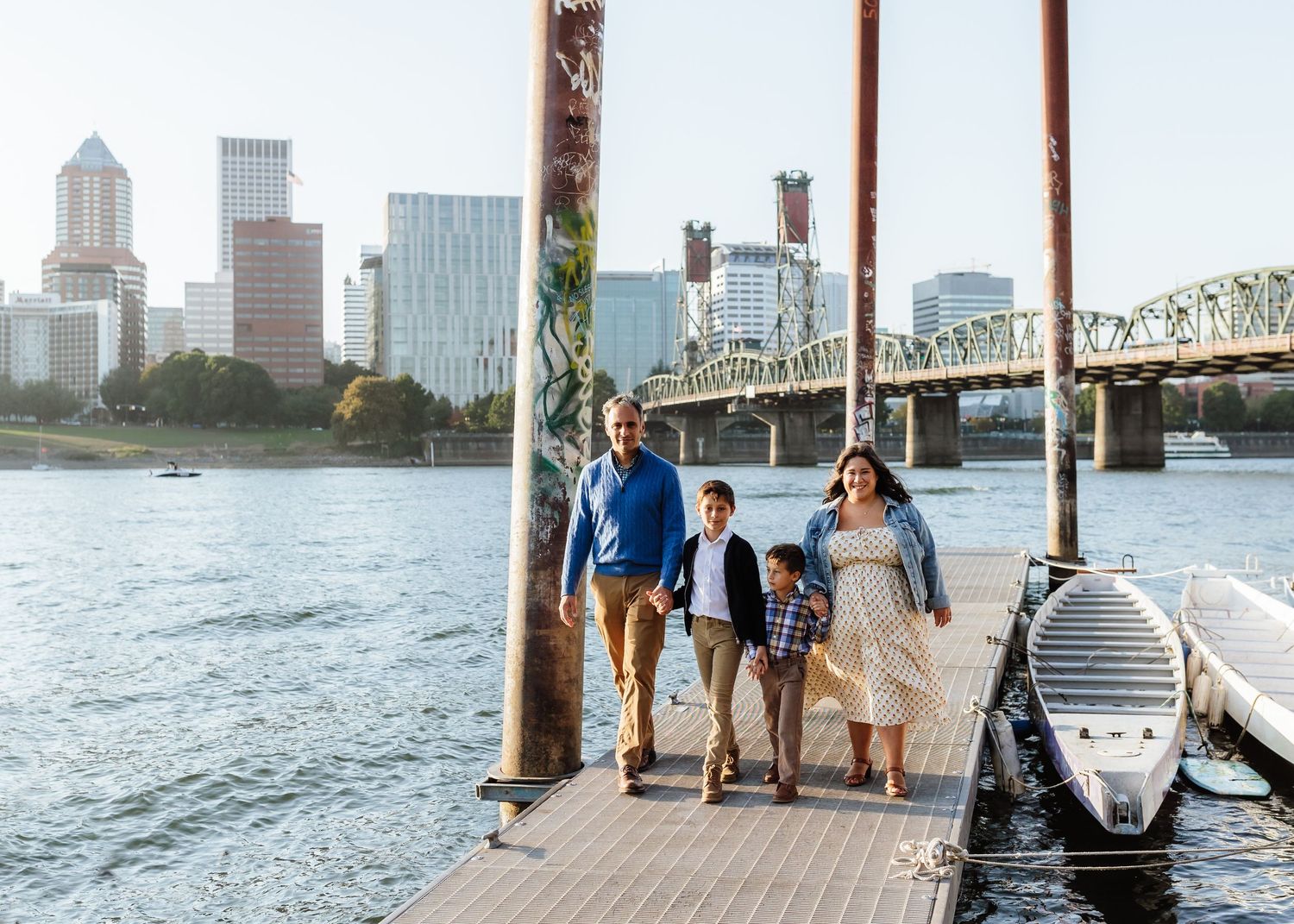 Family of four walking on a waterfront dock with a city skyline and bridge in the background on a sunny day.