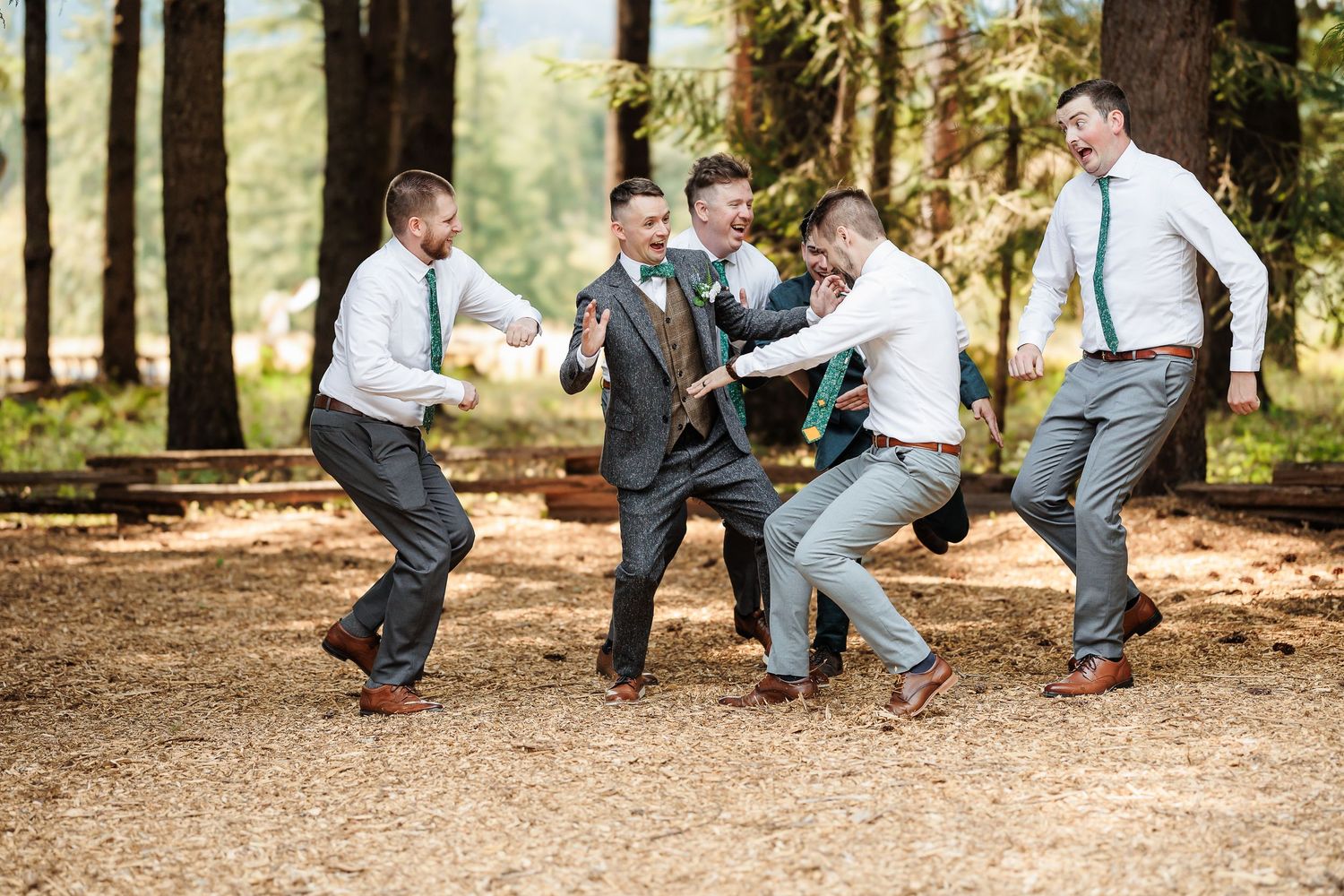 Groomsmen in white shirts and gray pants laugh and dance together in a sunny forest clearing.