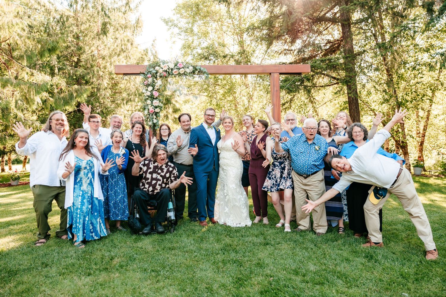Large wedding group celebrating with couple under wooden arch in sunny outdoor setting.