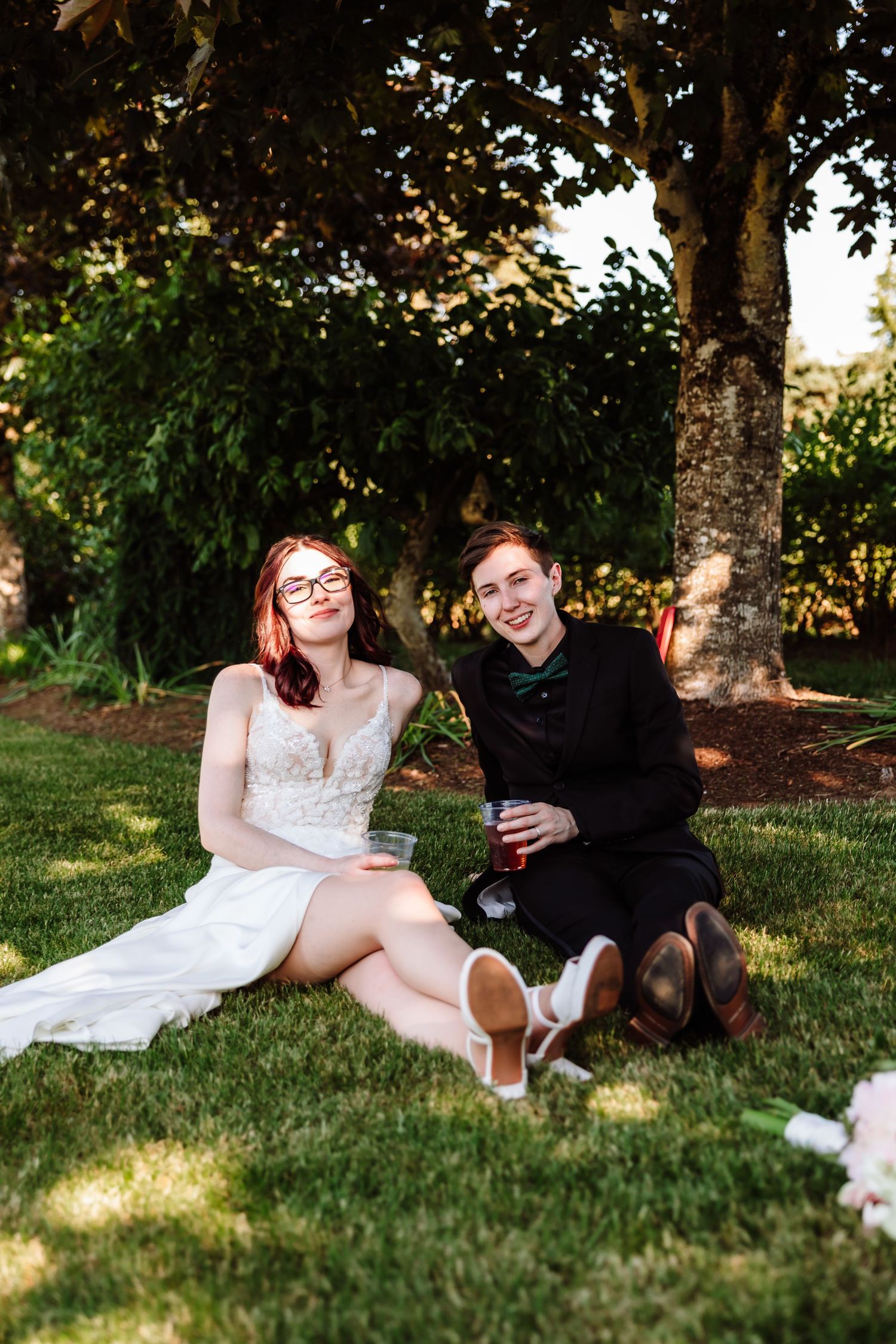 Newlywed couple sits relaxed on green grass under trees, holding drinks and smiling.