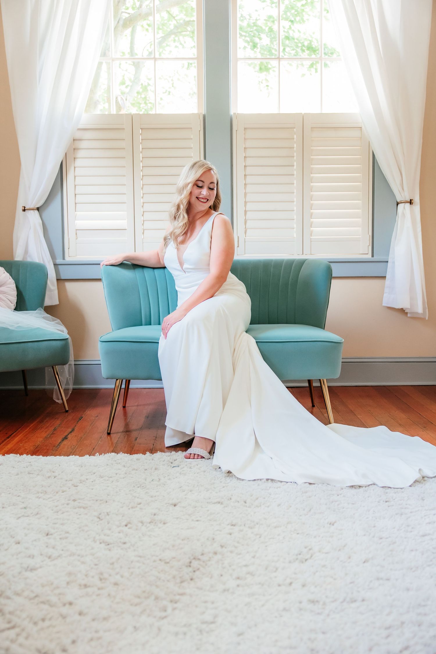Bride in a sleek white gown sitting gracefully on a teal velvet chair by window.