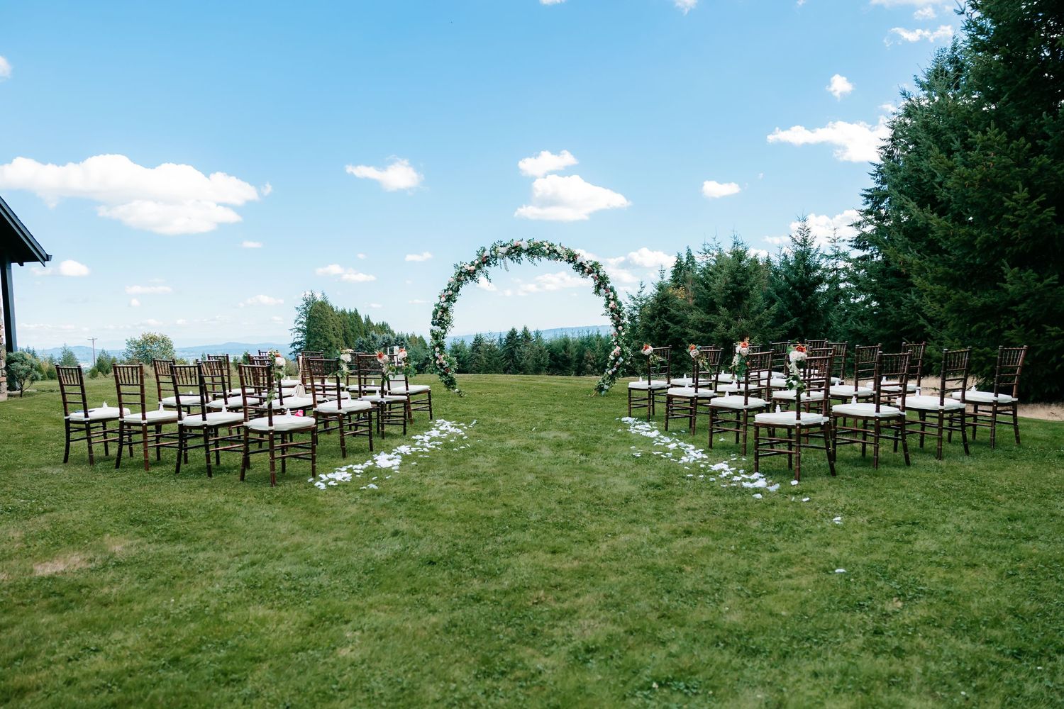 Elegant outdoor wedding ceremony setup with circular floral arch on a green hillside under blue sky.