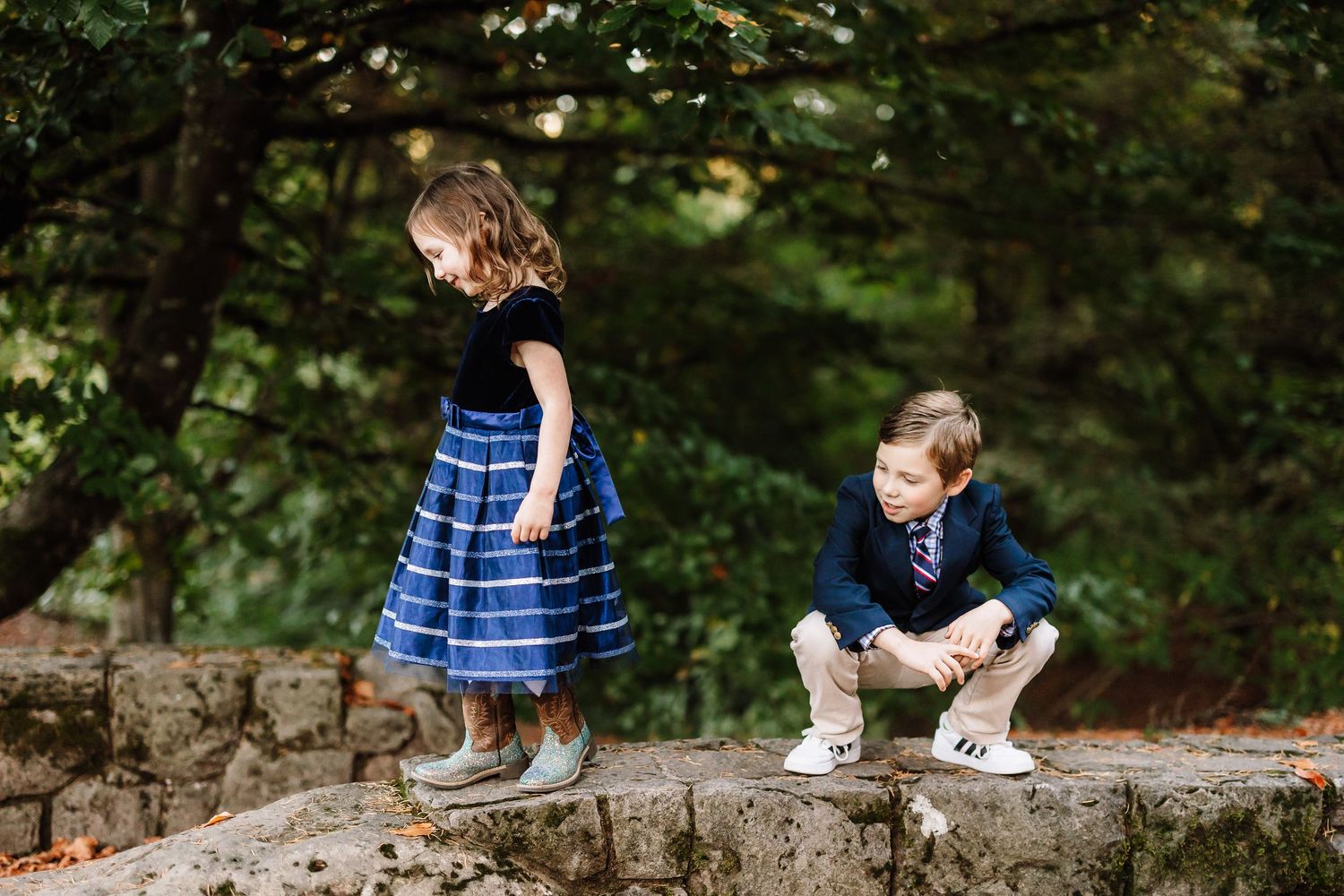 Two children in formal attire playing on a stone wall surrounded by lush green trees.