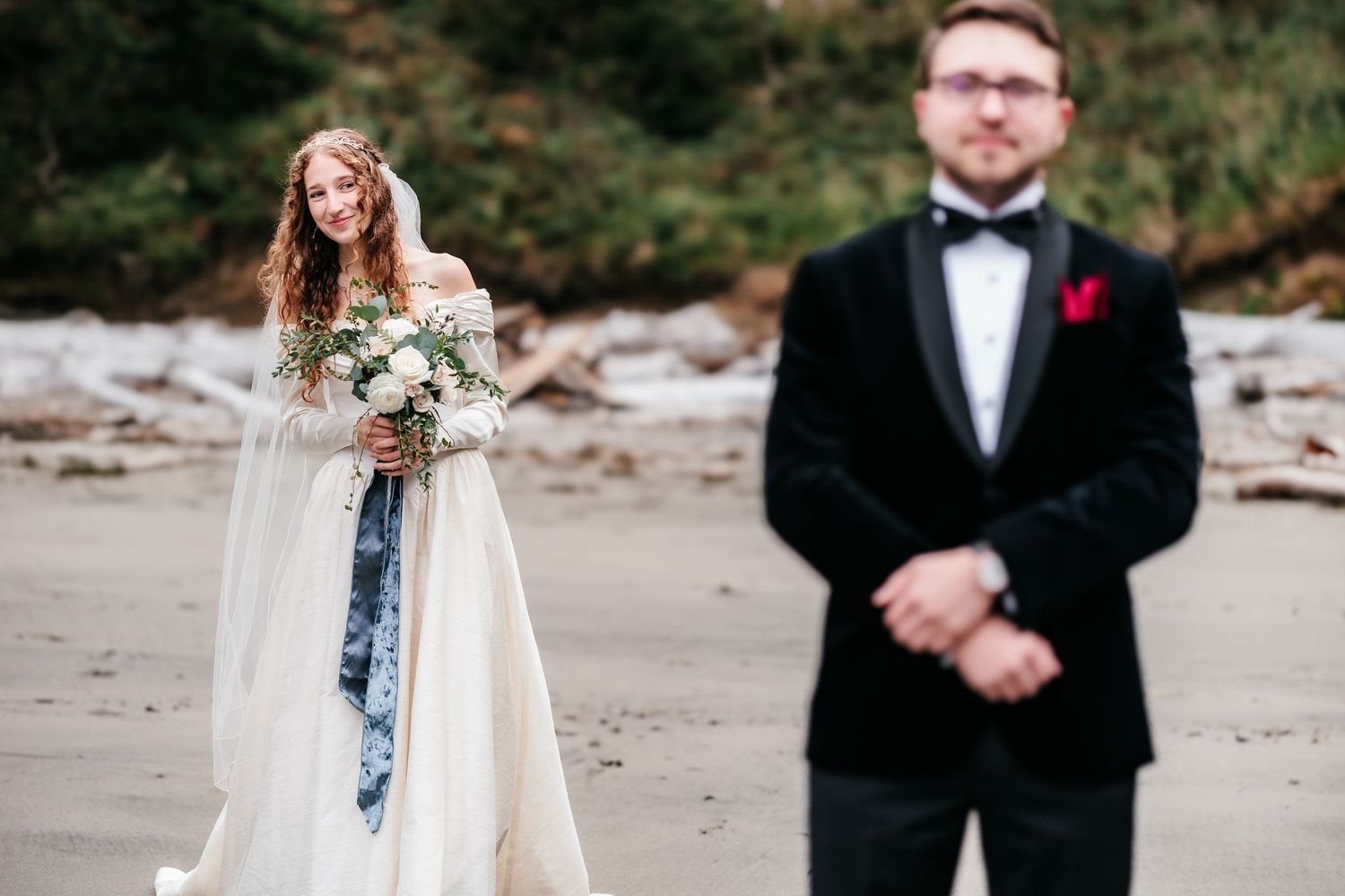 Bride smiles holding bouquet during beach first look with groom in tuxedo.