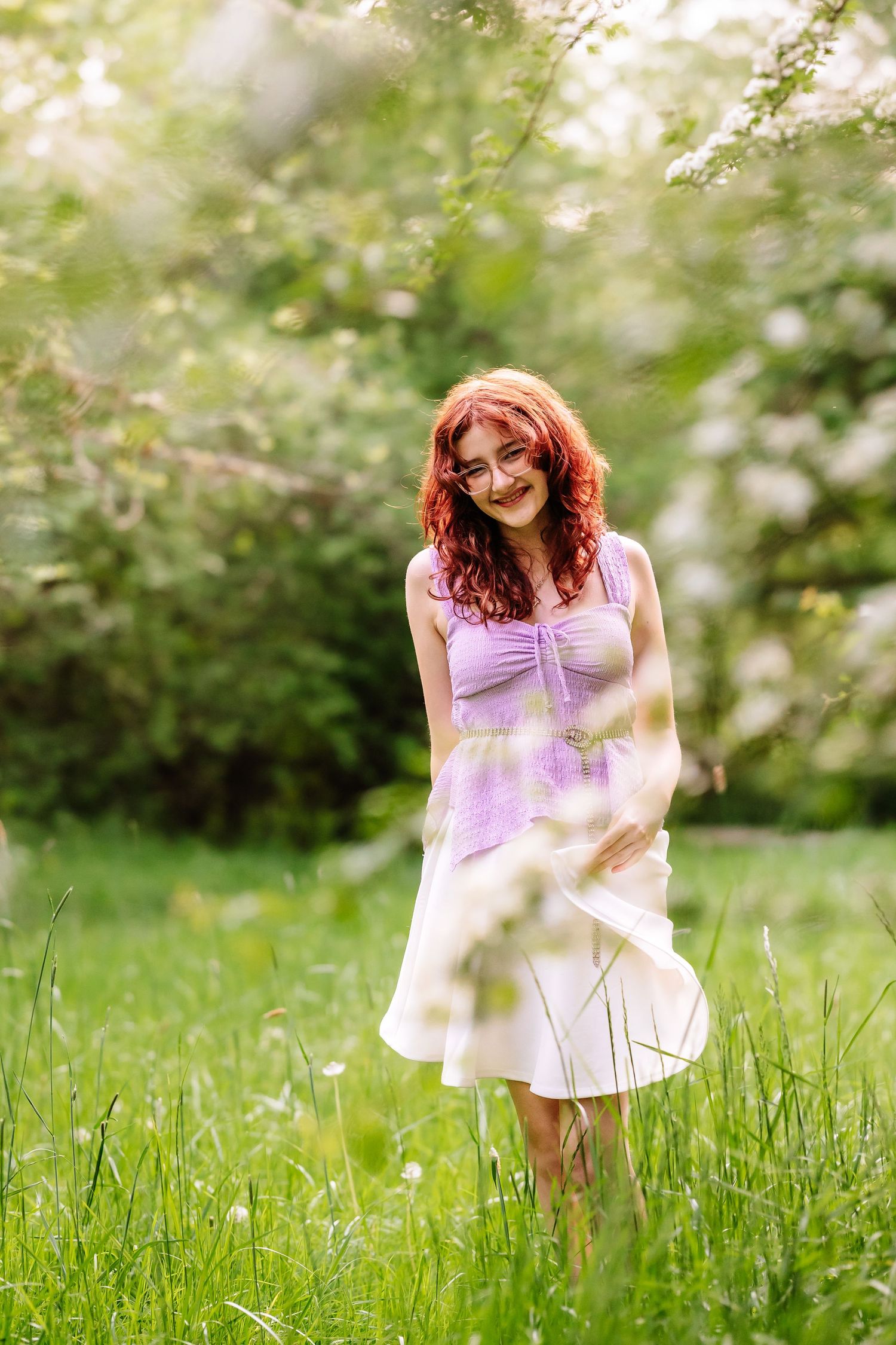 Smiling redhead woman in purple top and white skirt standing in lush green garden with blooming trees.