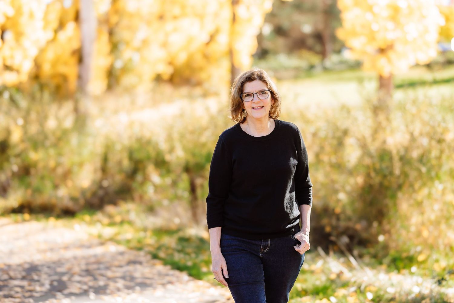 Woman in black sweater walking on a sunlit path surrounded by golden autumn foliage in a park.