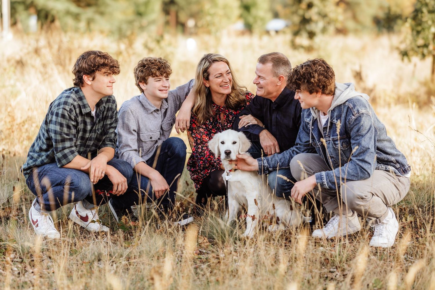 Family of five with a white puppy sitting together in a golden field, smiling and enjoying a warm outdoor photo session.