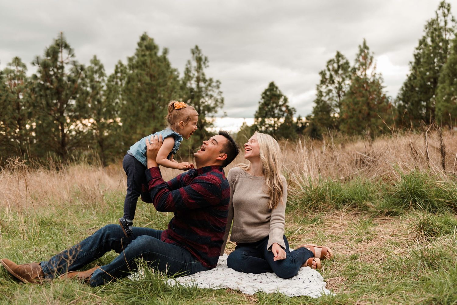 Family of three sitting on blanket in field, parents smiling as toddler girl stands between them outdoors.
