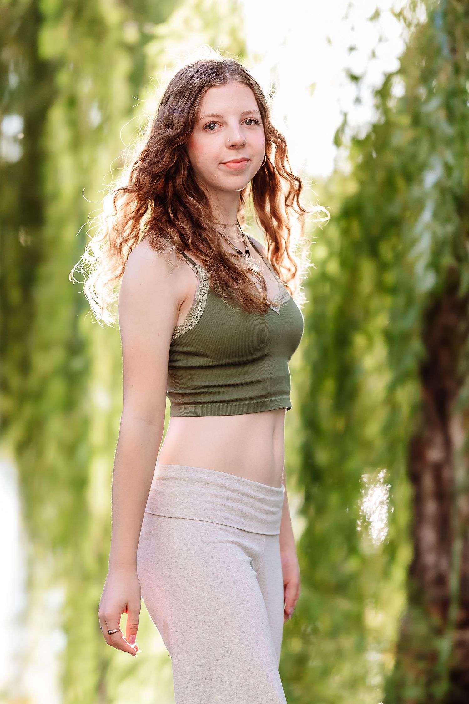Young woman with curly hair wearing a green crop top and gray sweatpants posing outdoors near willow trees.