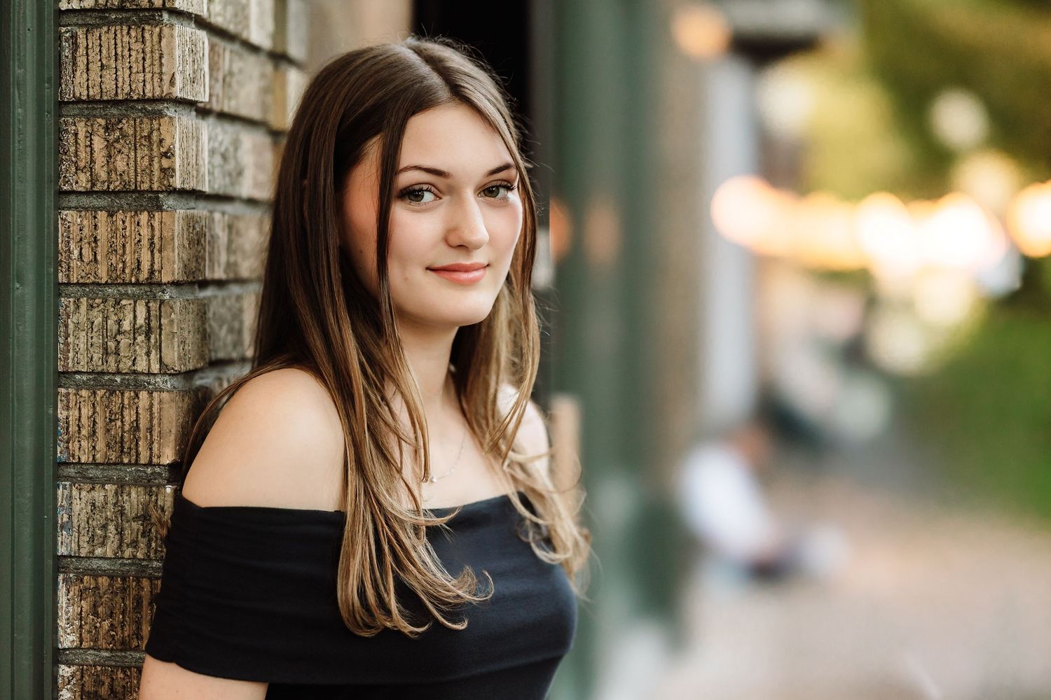 Young woman with long brown hair in black off-shoulder top leaning against brick wall outdoors.