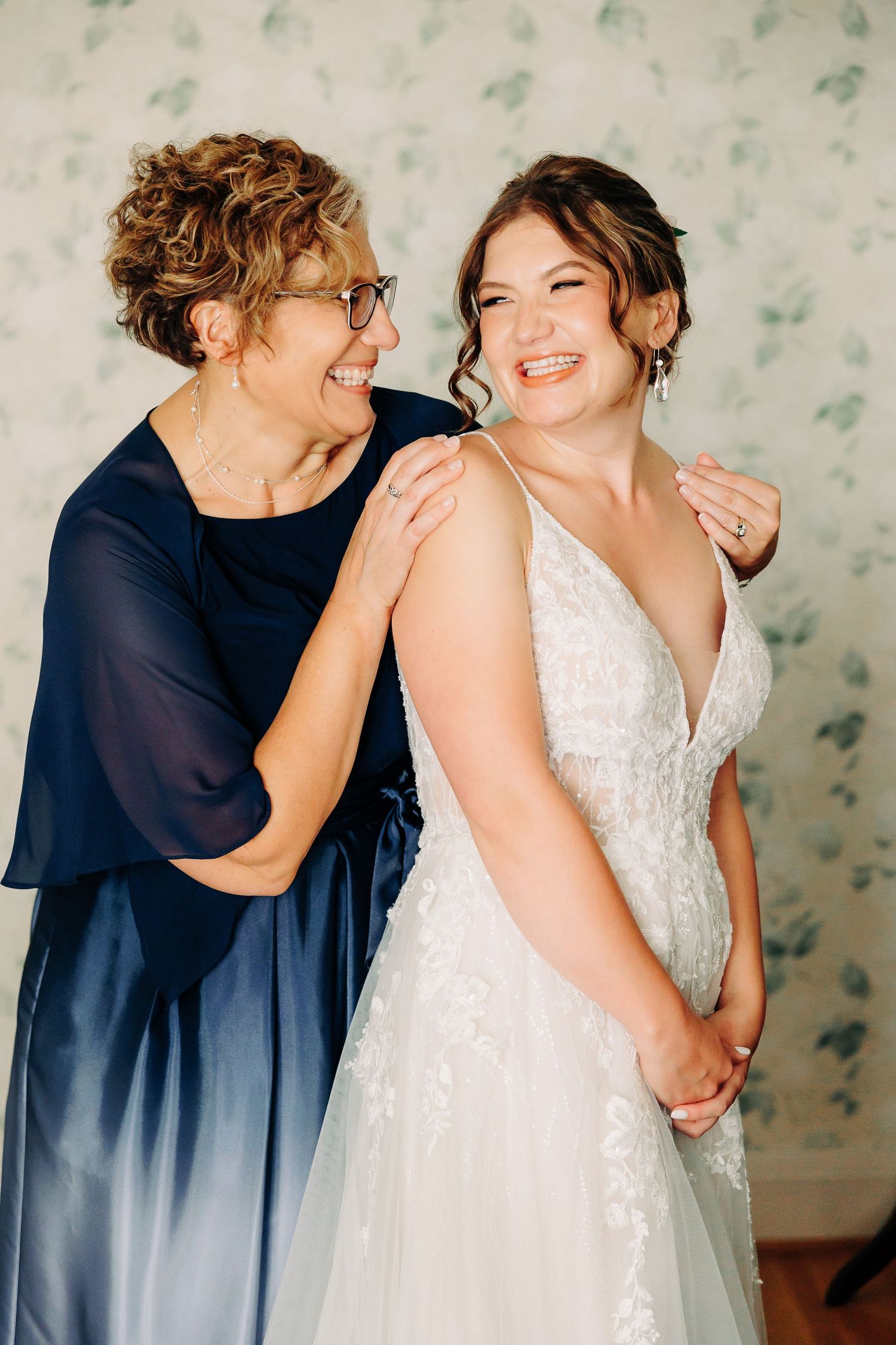 Bride laughing joyfully with older woman in navy dress during wedding day preparations.