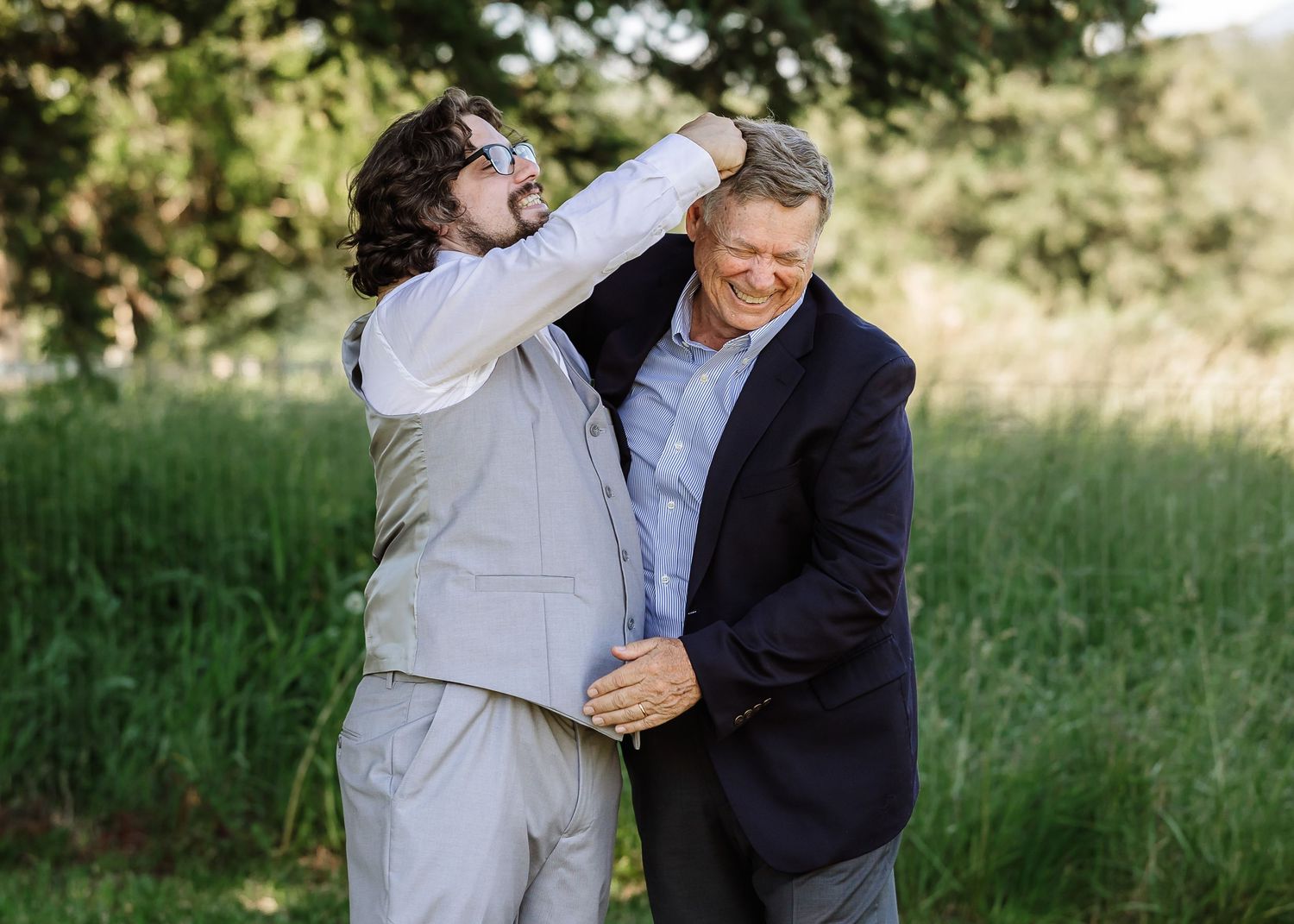 Groom in gray vest playfully adjusts older mans tie while both laugh in green field.
