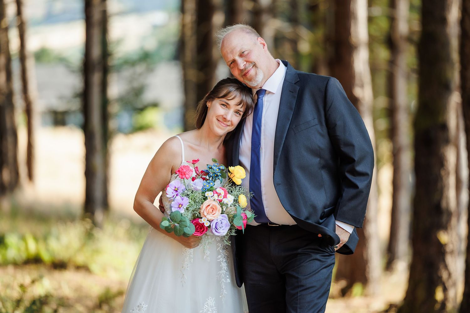 Bride holding colorful bouquet leans on groom in navy suit, smiling together in a sunlit forest setting.