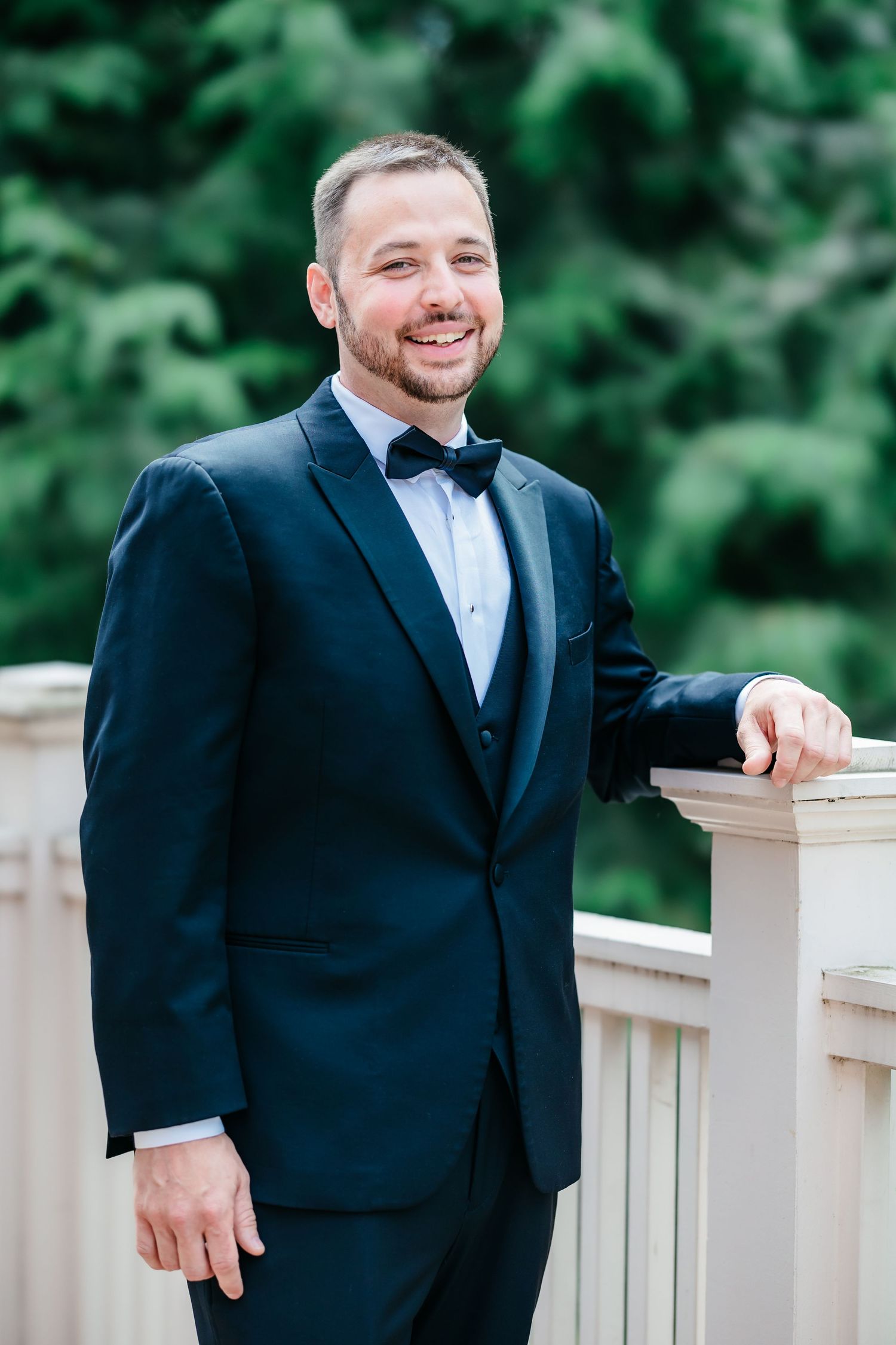 Smiling groom in a black tuxedo with bow tie leaning on a white railing with green trees behind him.