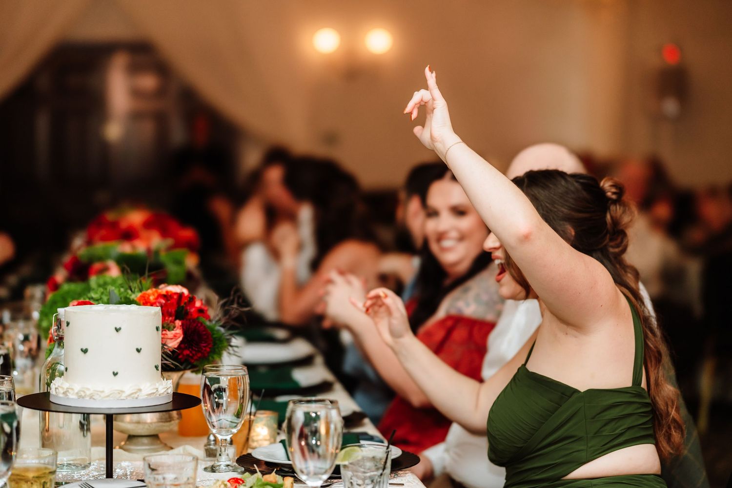Wedding guest in green dress raises hand excitedly at elegant reception dinner table with white cake.