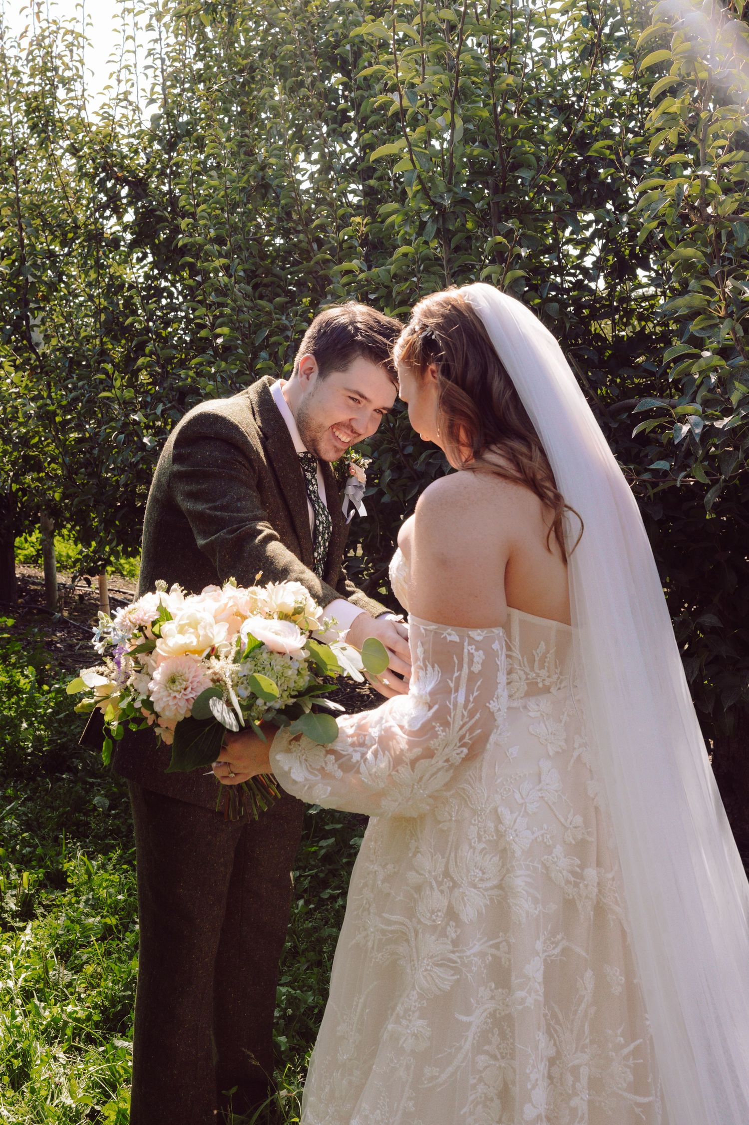 Bride and groom touching foreheads and smiling while sharing bouquet in sunny orchard.