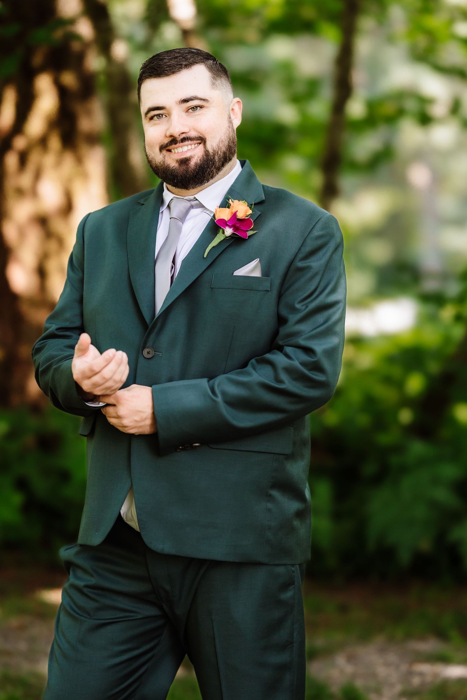 Smiling bearded groom in dark green suit with colorful boutonniere poses outdoors in garden setting.