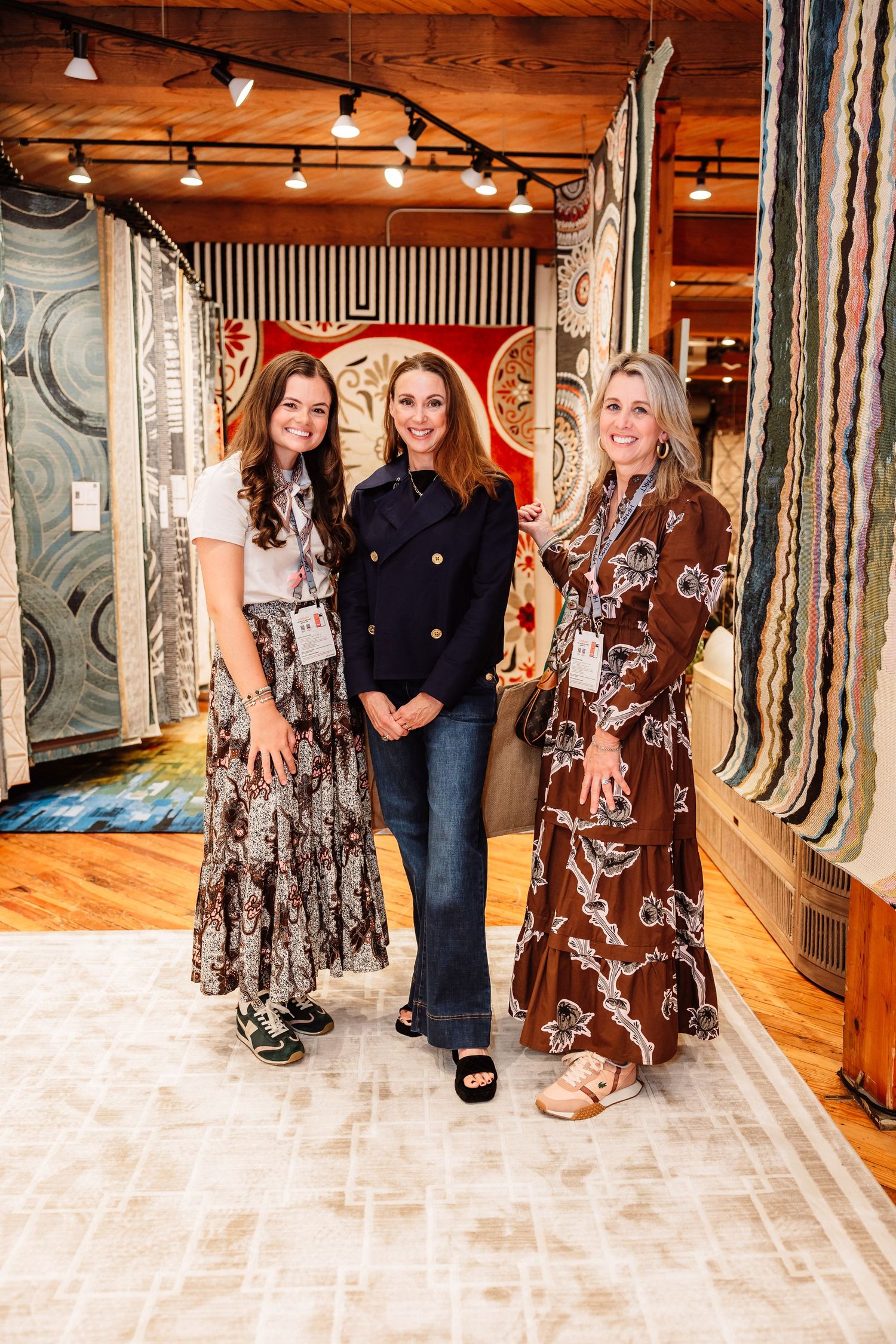 Three women smiling in a rug showroom surrounded by colorful patterned rugs on display.
