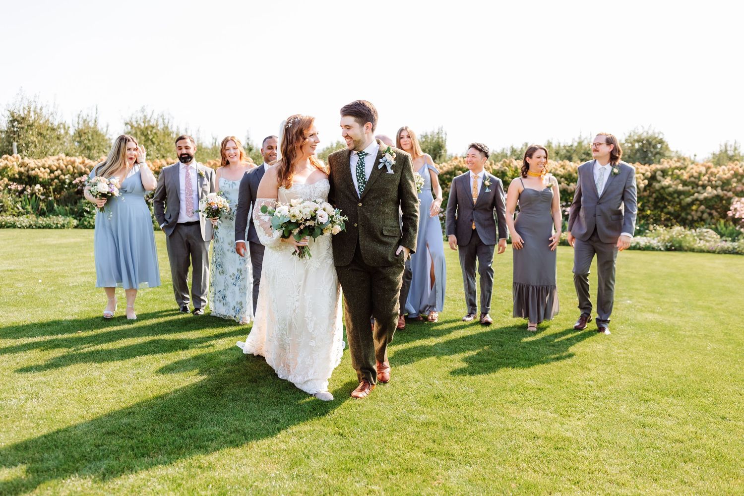 Bride and groom walk with their wedding party across a lush green lawn on a sunny day.