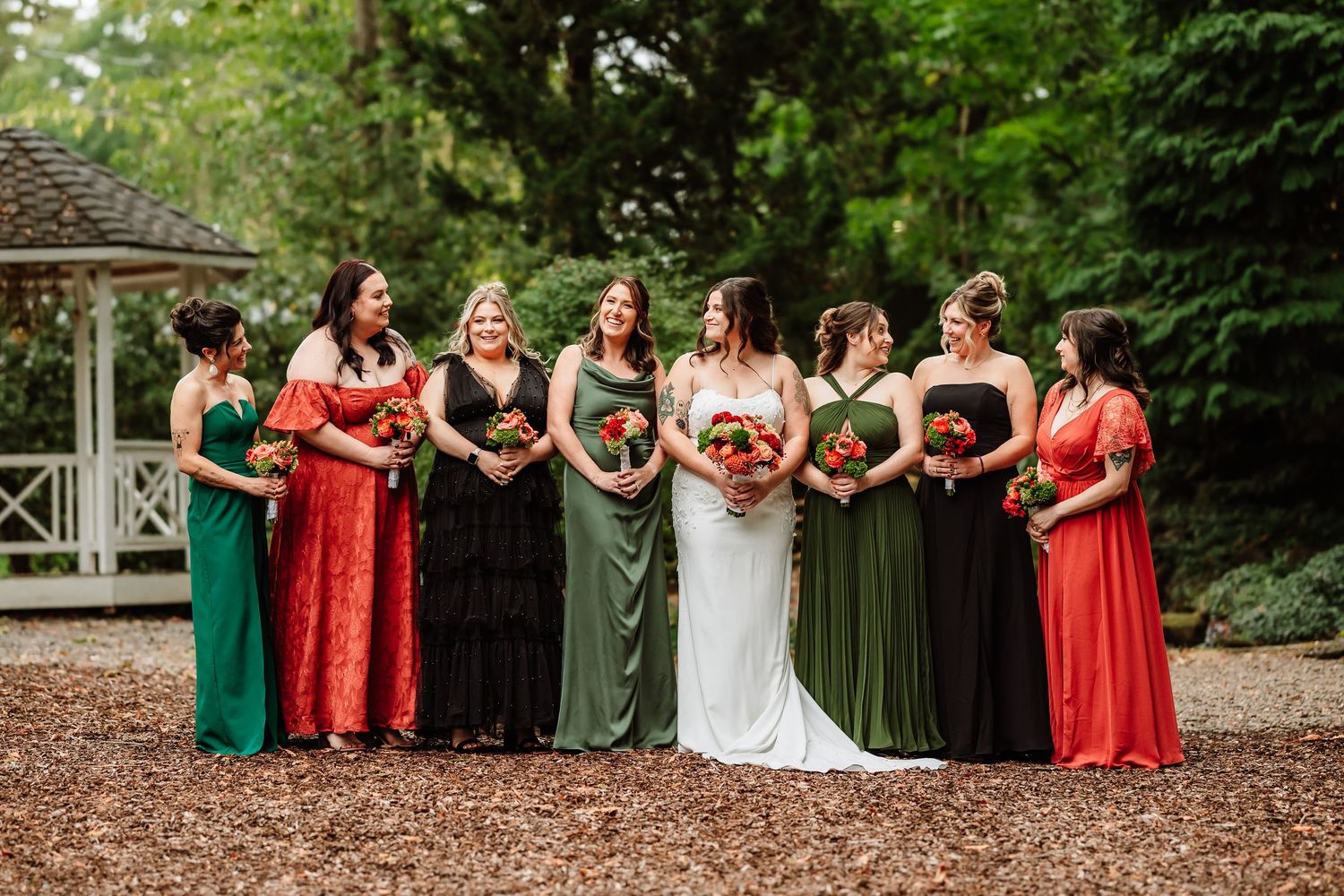 Bridesmaids in mixed red, green, and black gowns hold colorful bouquets alongside a bride in white outdoors.