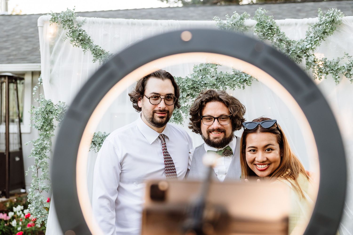 Three smiling wedding guests pose together viewed through a ring light photo booth frame.