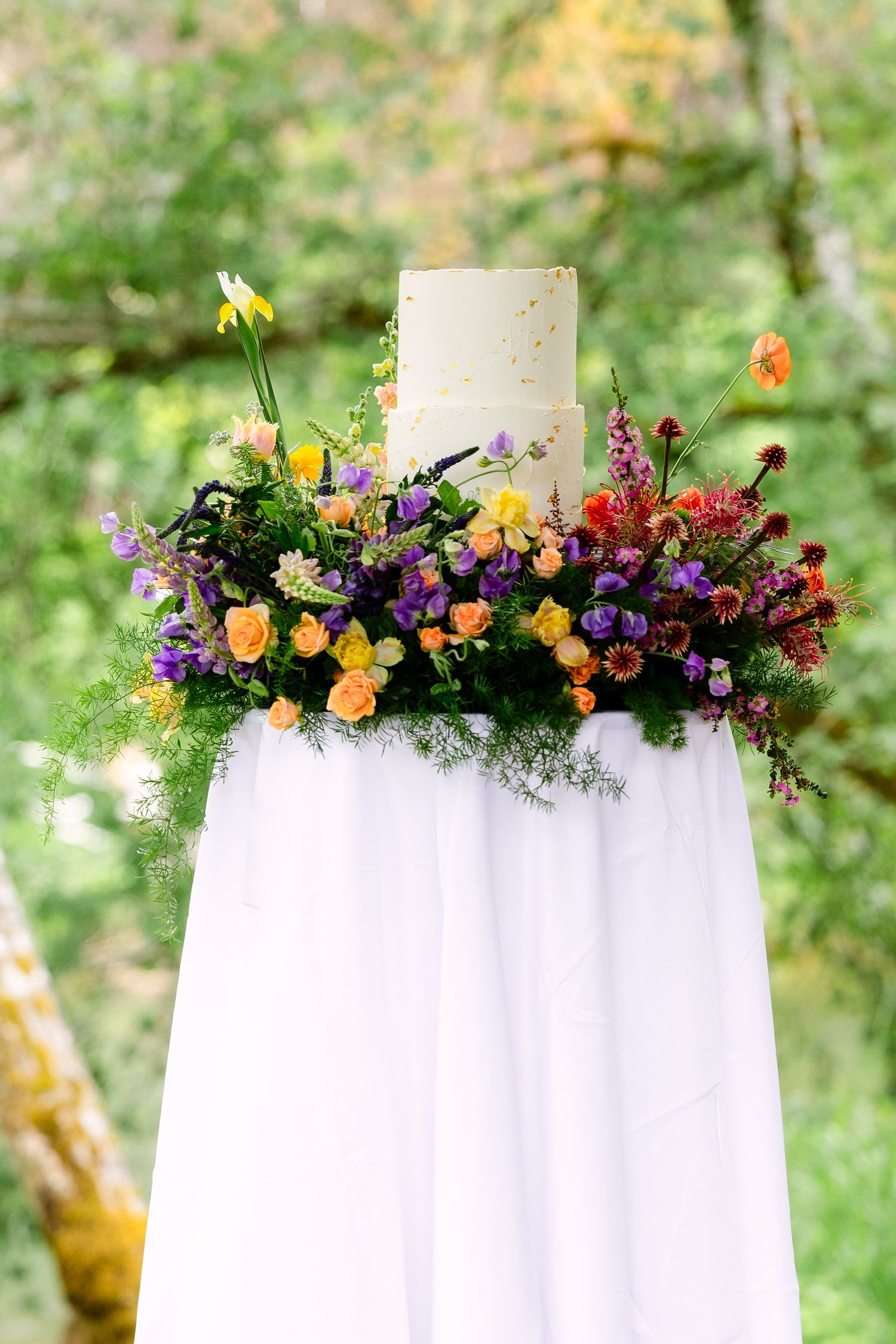 Two-tier white wedding cake surrounded by colorful wildflower arrangement on a white draped table outdoors.