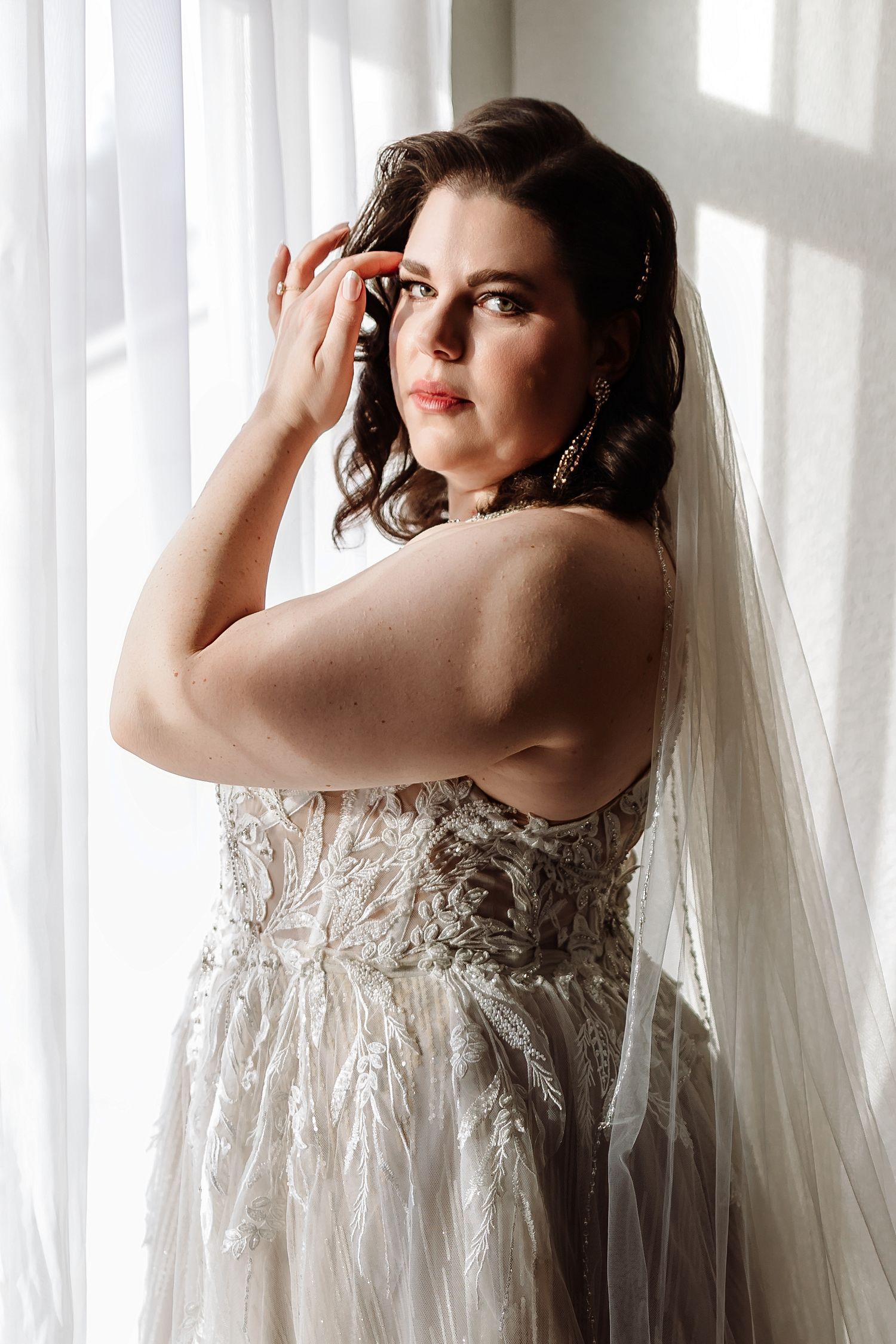 Elegant bride in lace wedding dress with veil posing by a bright window.