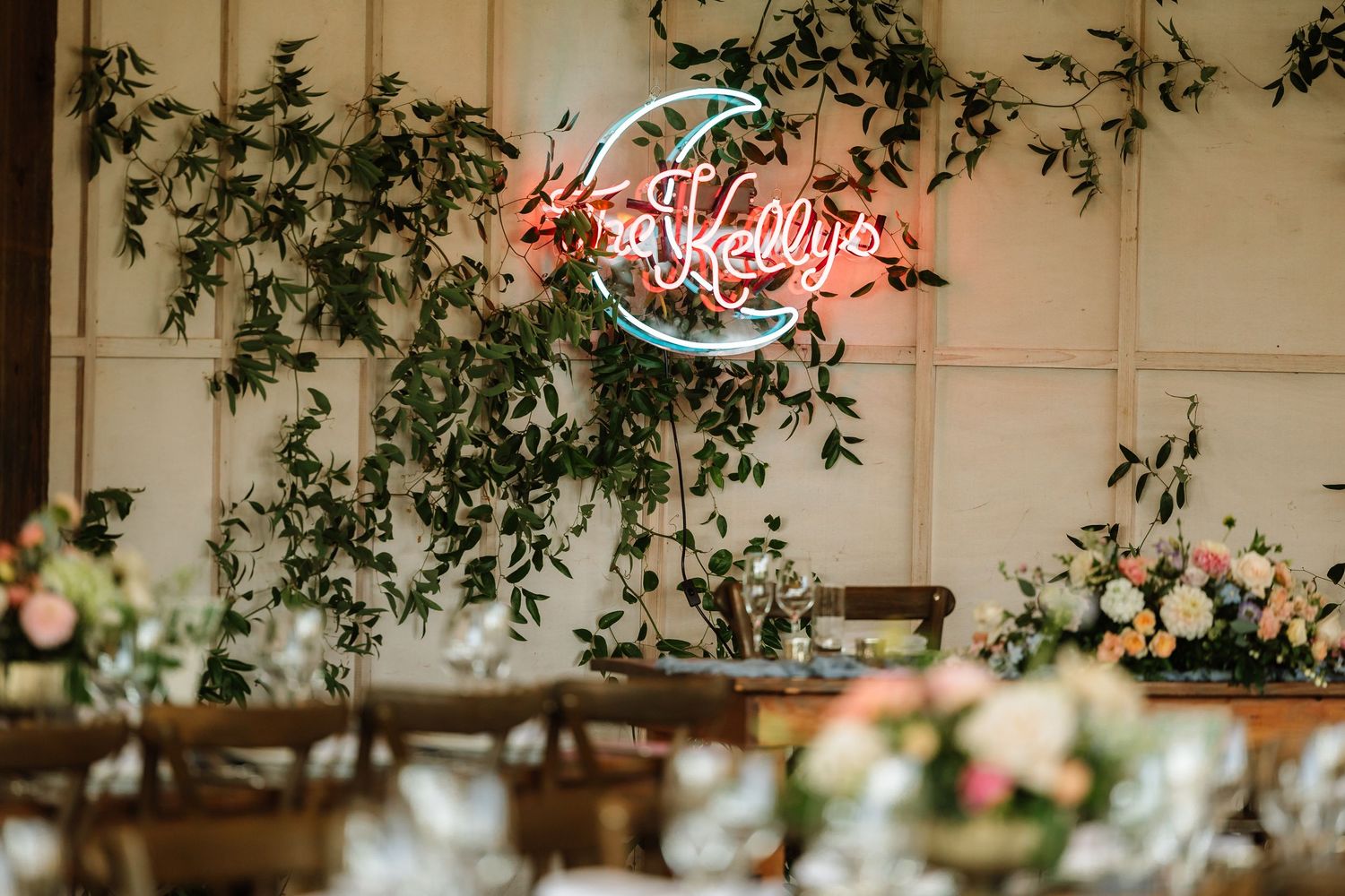 Glowing pink neon sign reading Kellys above lush greenery wall decor at elegant wedding reception.