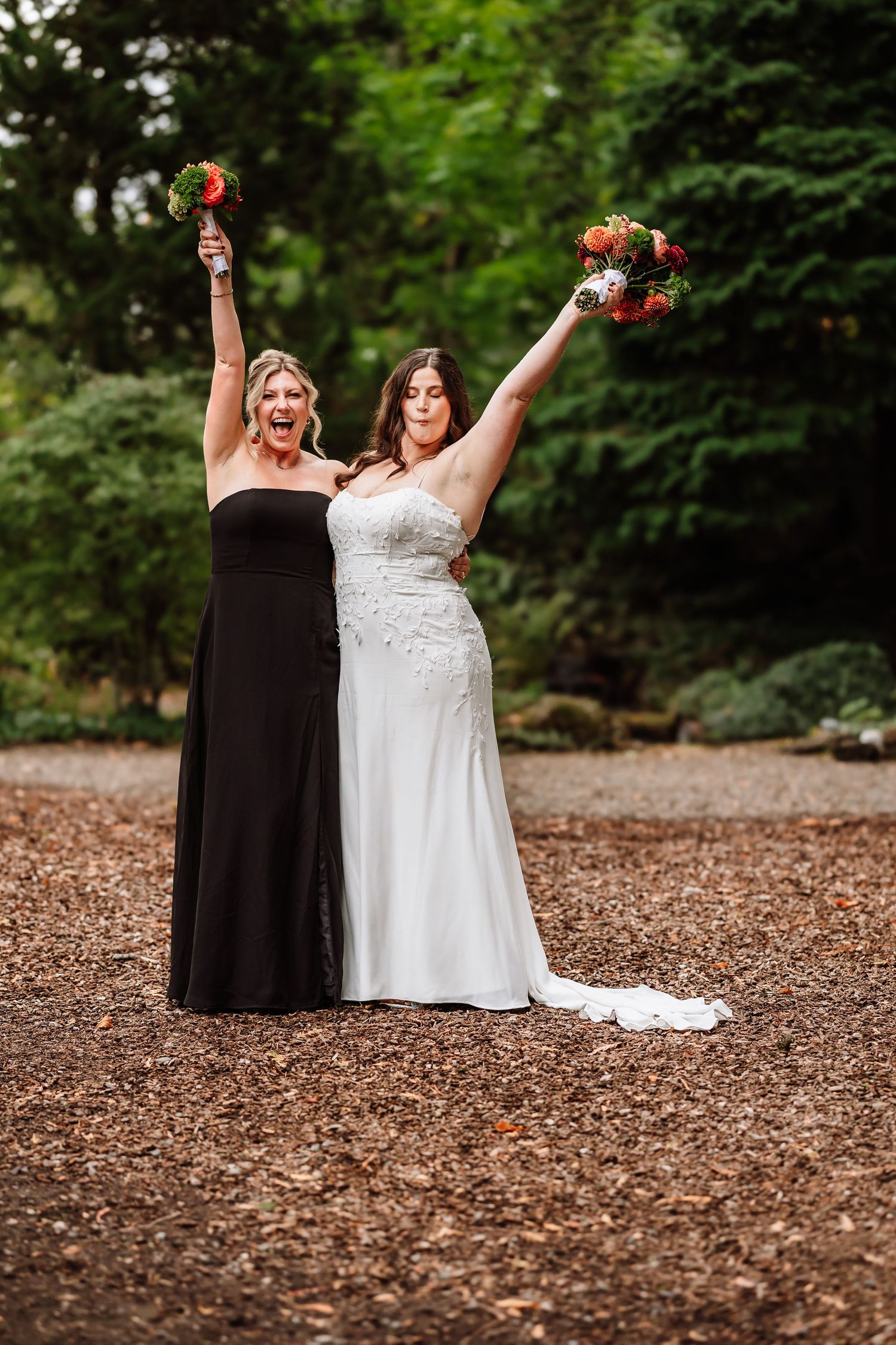 Two women celebrate joyfully raising bouquets of orange flowers in a garden, one in a wedding dress.