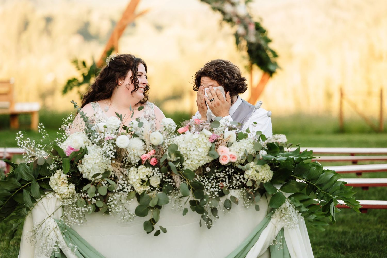 Bride and groom share a laugh at floral-decorated sweetheart table during outdoor wedding reception.
